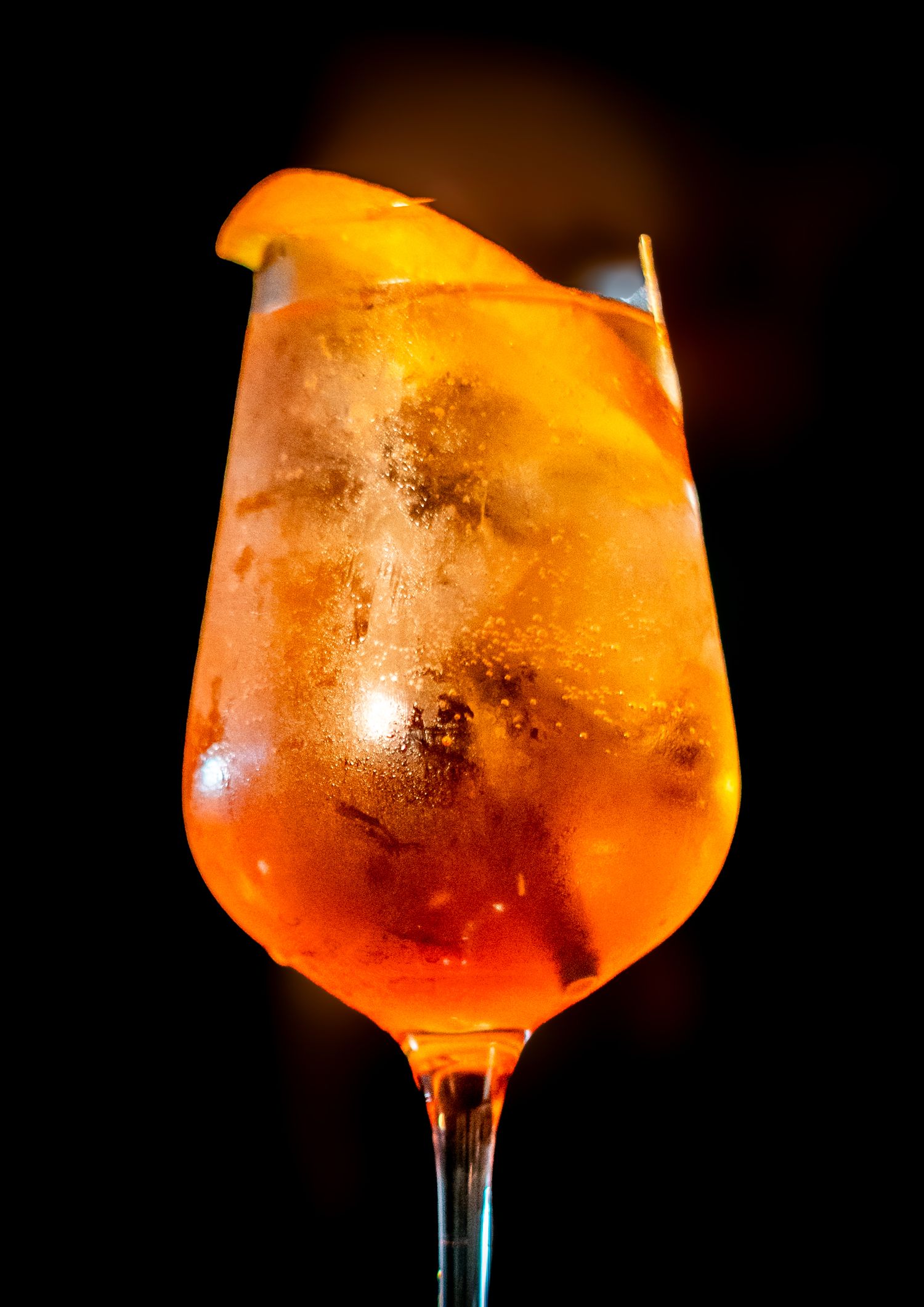 Close-up of an orange-colored cocktail with ice in a stemmed glass against dark background.