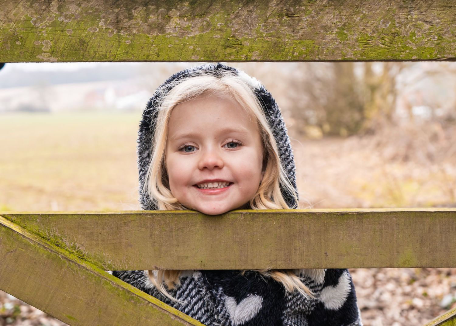 A young person peeks through wooden fence slats while wearing a patterned winter coat.