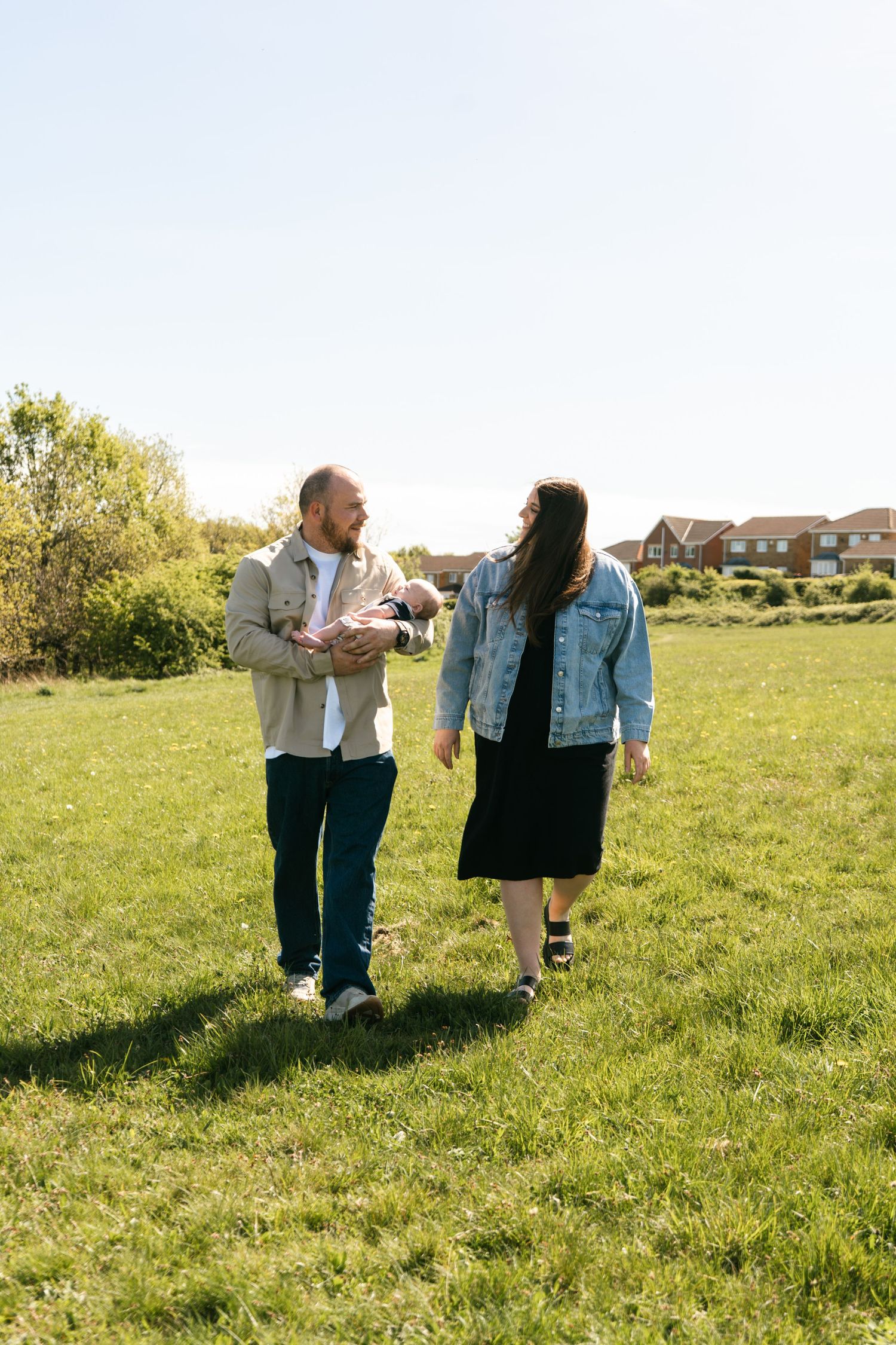 A couple walks together on a sunny grassy field with houses visible in the background.