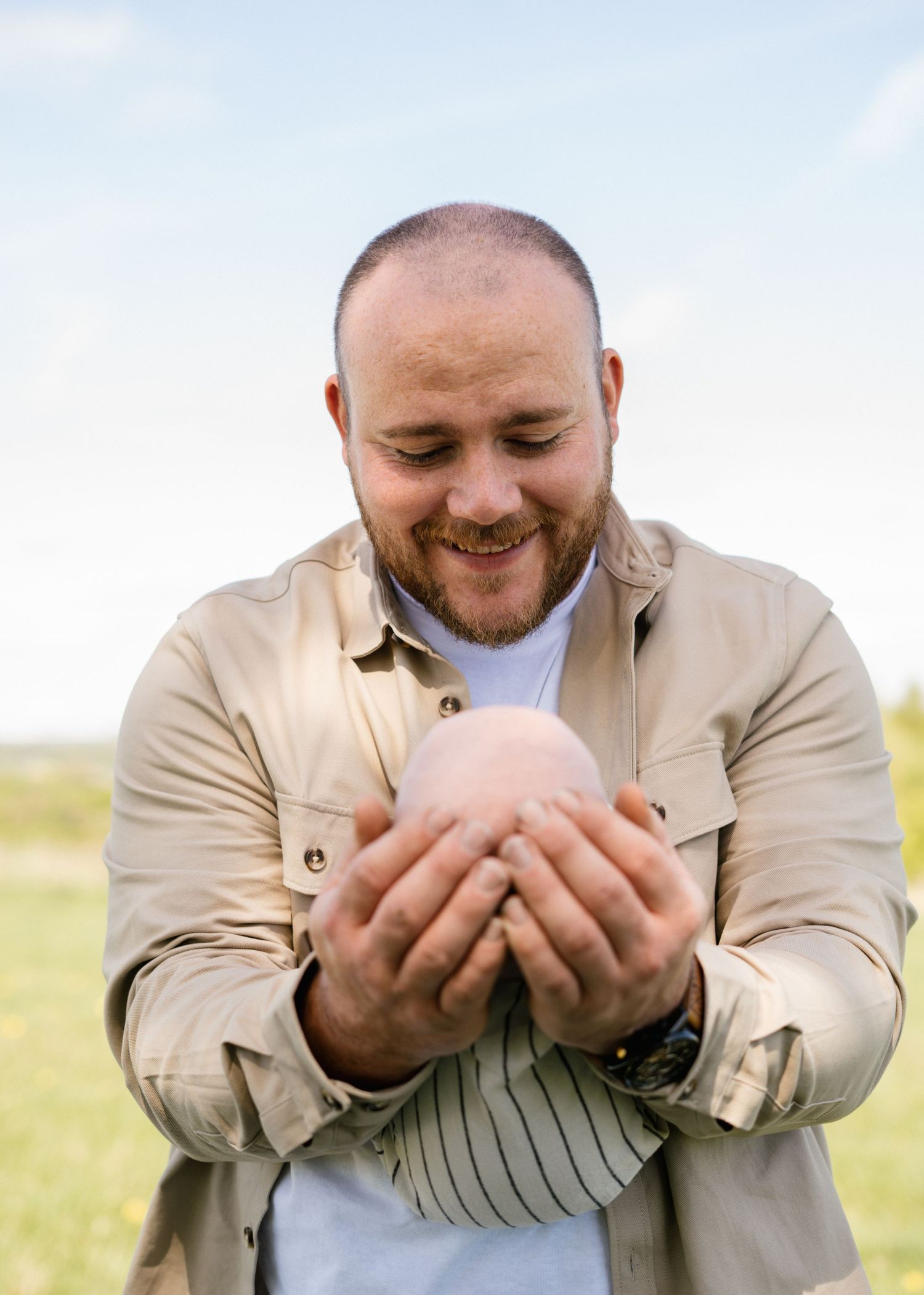 A person in a beige jacket smiles while holding something precious in their hands outdoors.