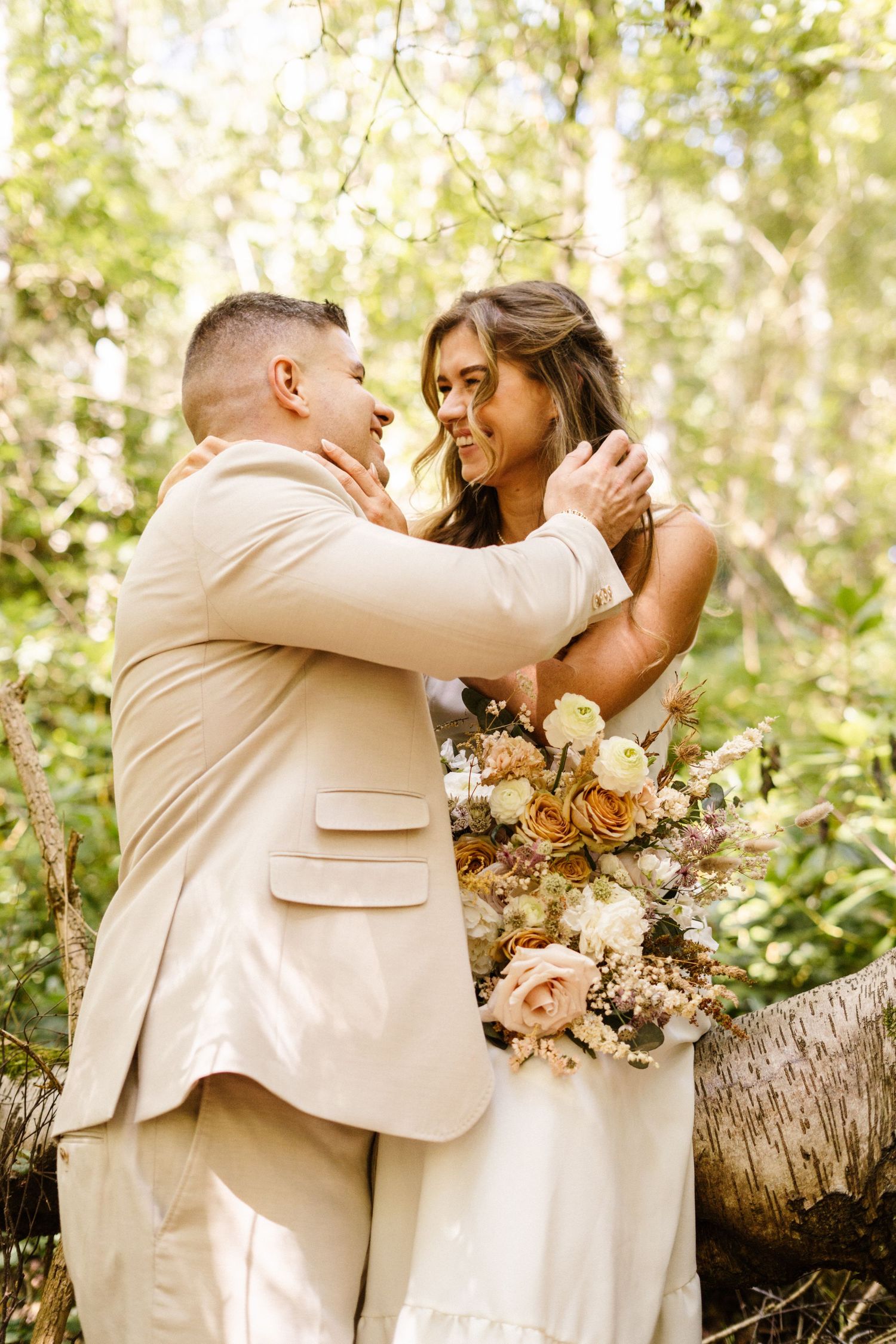 Couple in light colored wedding attire embrace while holding floral bouquet in a sunlit garden.