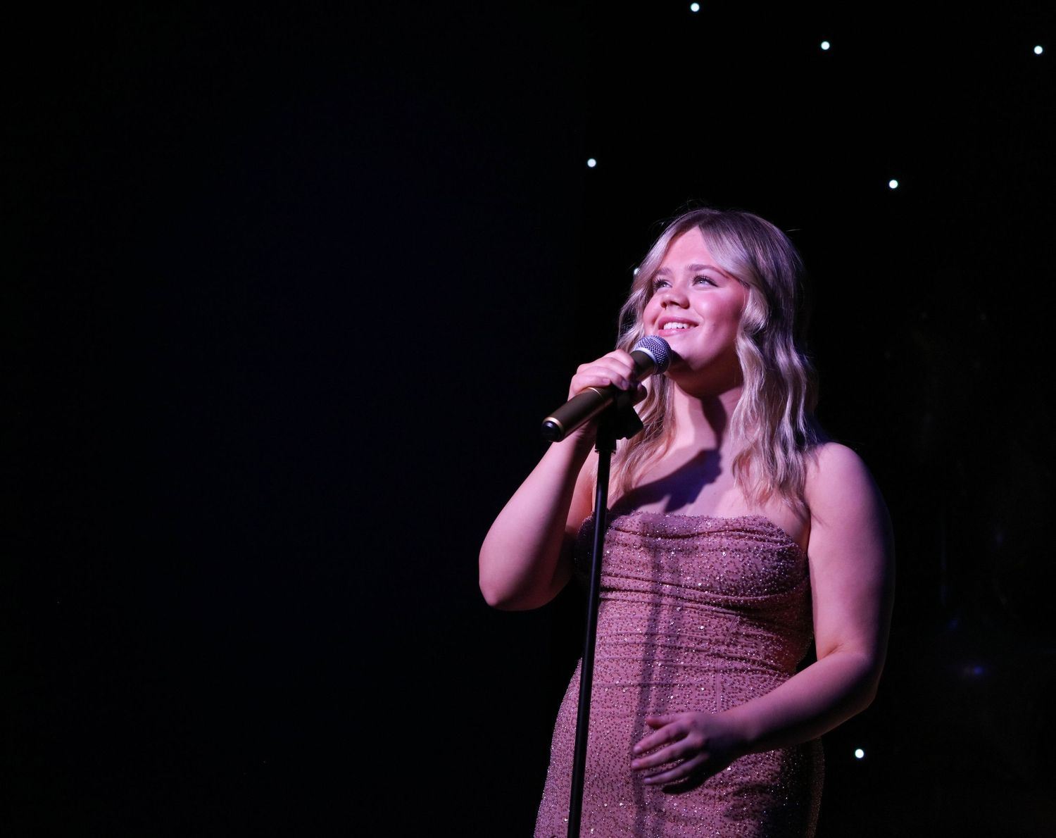 Performer in sequined dress sings emotively against dark stage with twinkling lights.