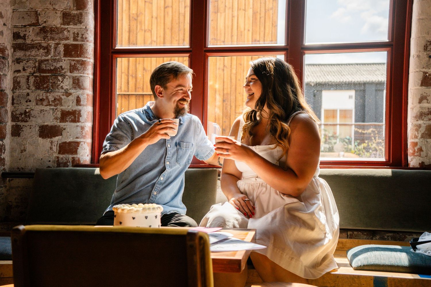 A couple sits together in a rustic brick room with windows.