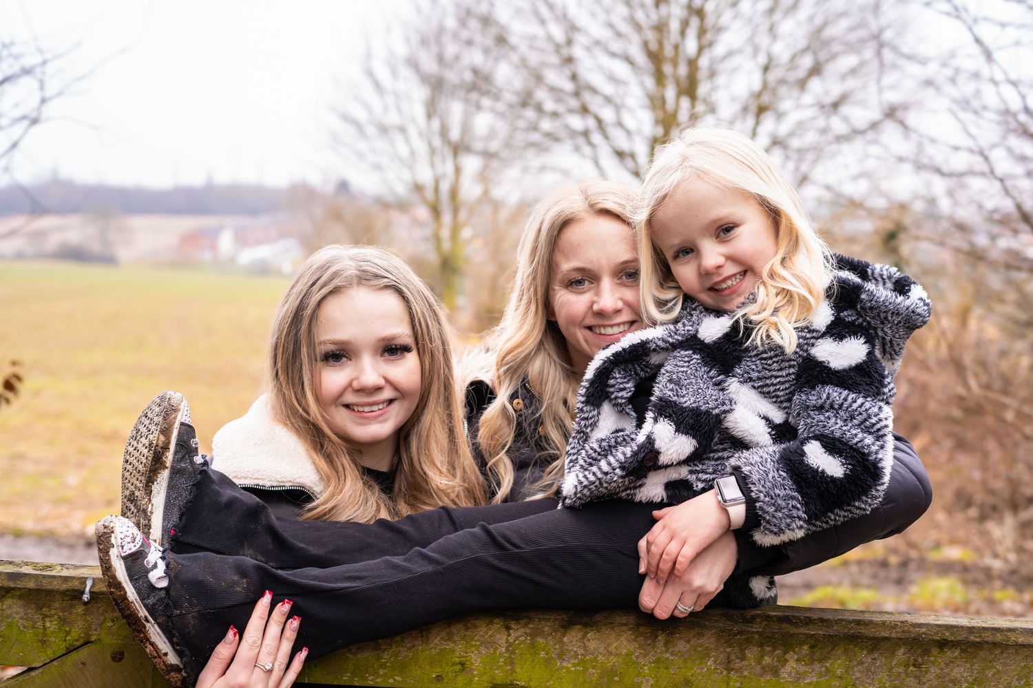 Three people share a joyful moment while sitting on a wooden gate in a rural setting.