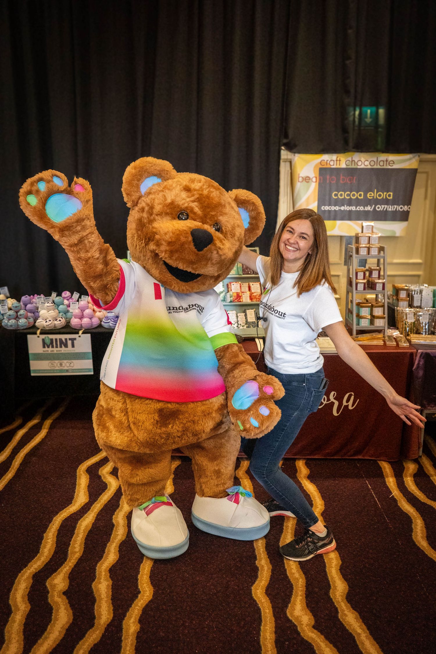 Large plush mascot character interacting playfully with attendees at an indoor event.