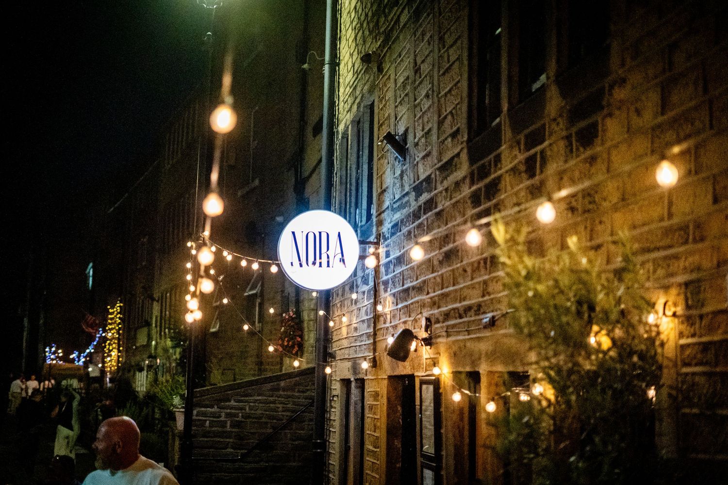 Illuminated NORA sign on brick wall with string lights creating ambient evening atmosphere.