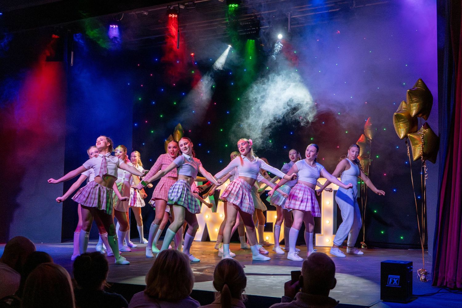 Dance ensemble performs choreographed number under colorful stage lighting with fog effects.