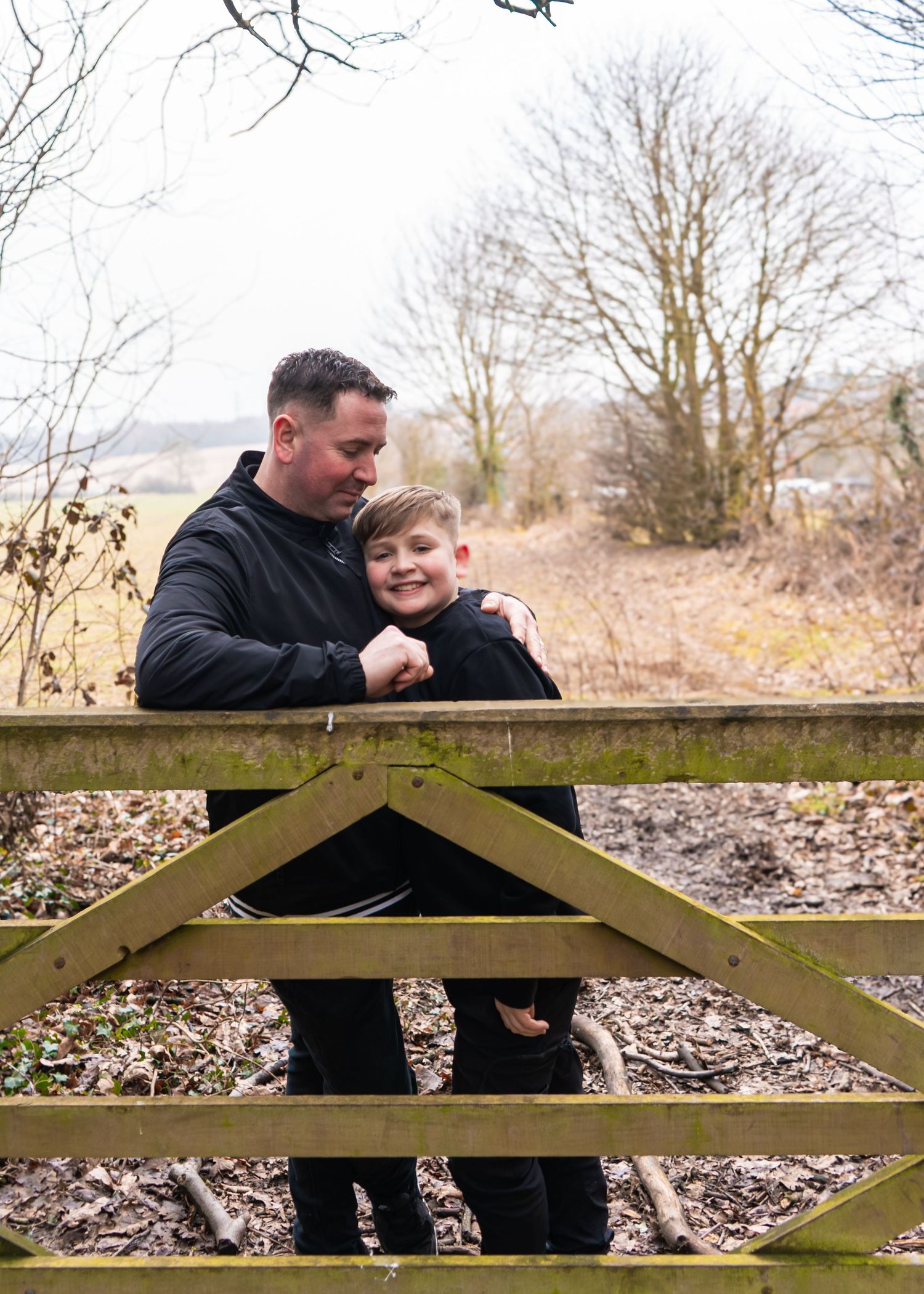 Two people in black jackets stand behind a rustic wooden gate on a winter day.