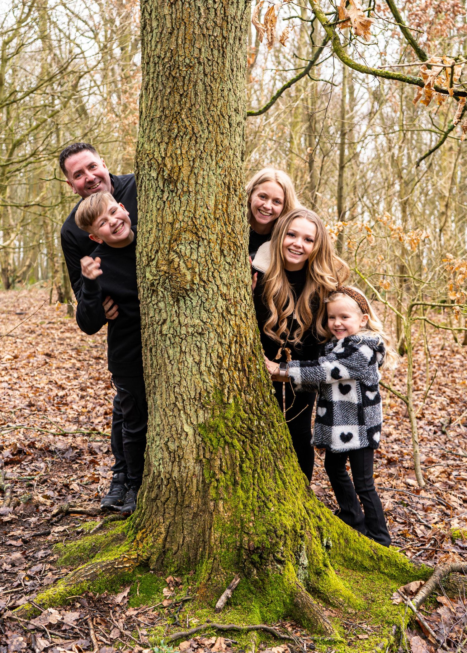 Fun family portraits with people hiding and peeking from behind a large tree in the woods.
