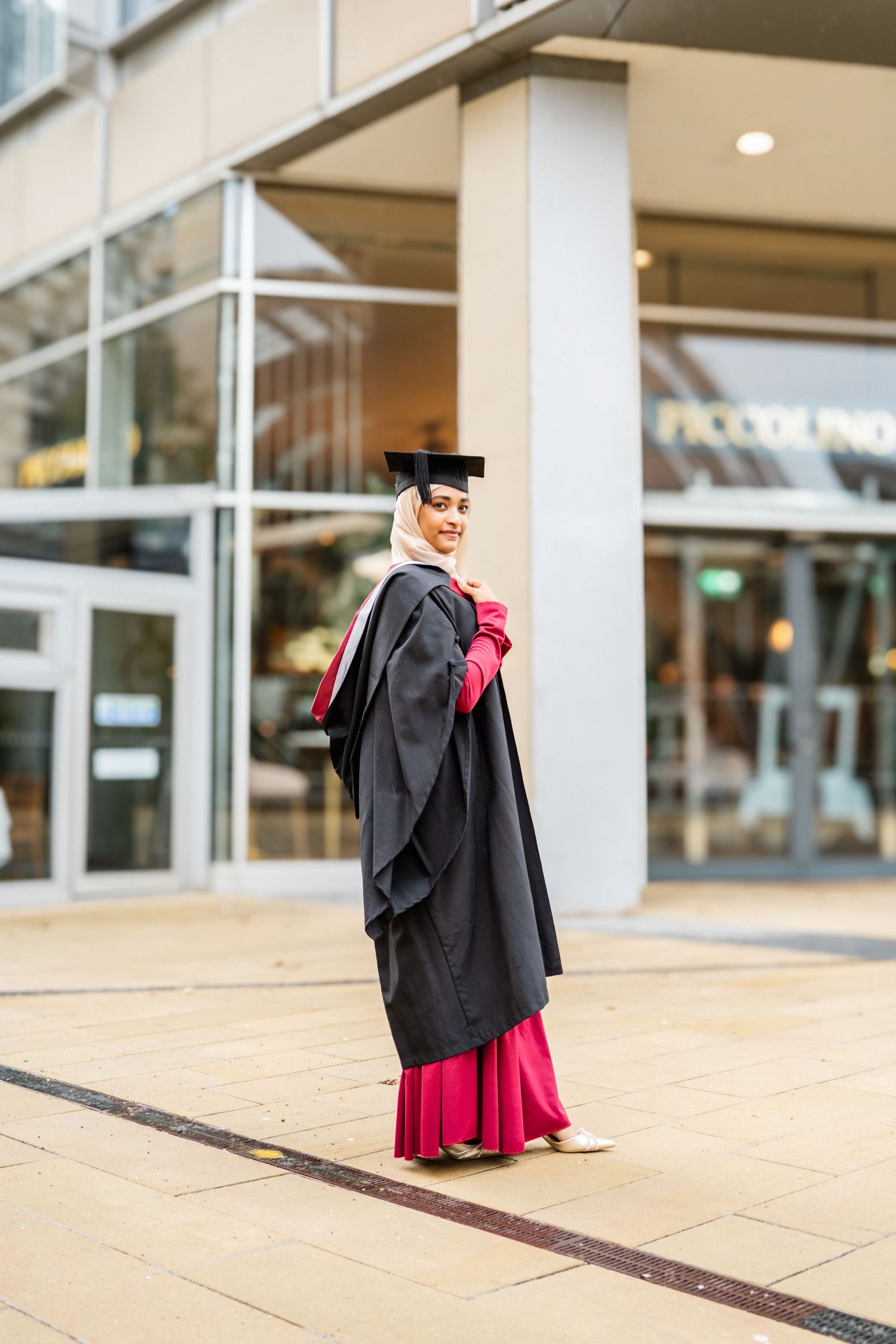 Graduate in black gown and red dress poses in front of modern building columns.