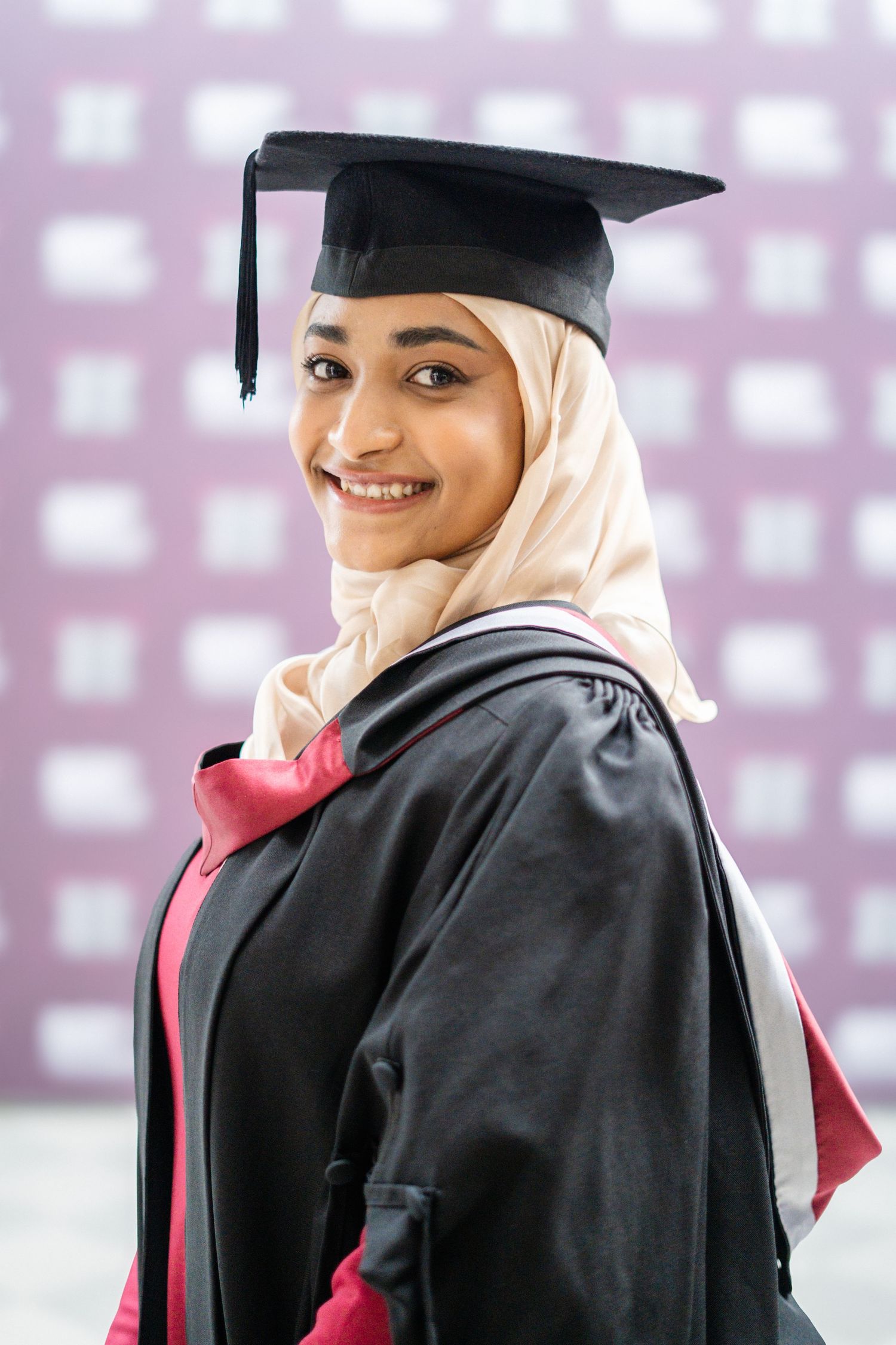 Graduate in academic regalia and hijab poses against purple patterned backdrop.