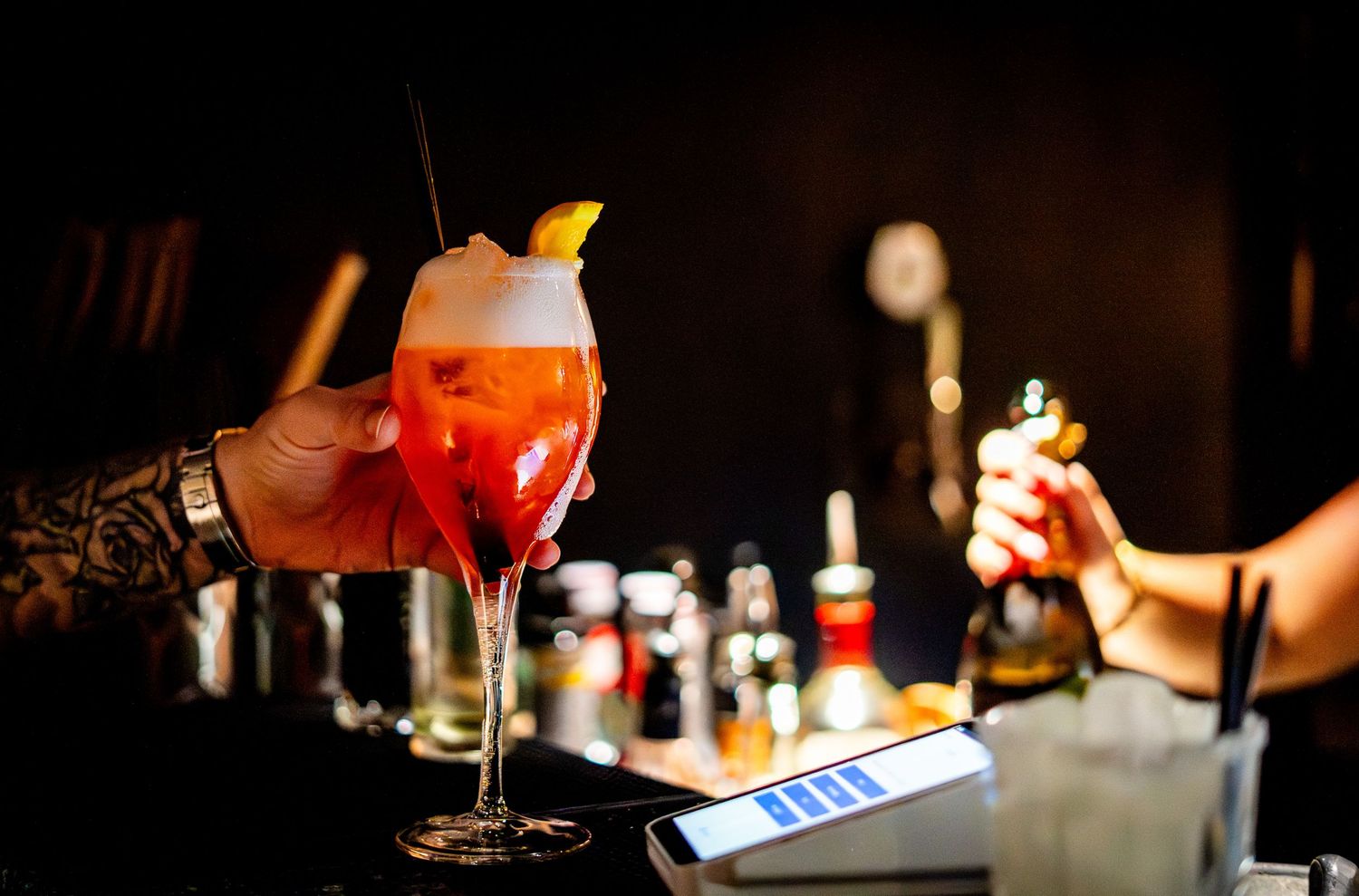 Colorful cocktails being prepared at a bar with bottles and payment terminal visible.