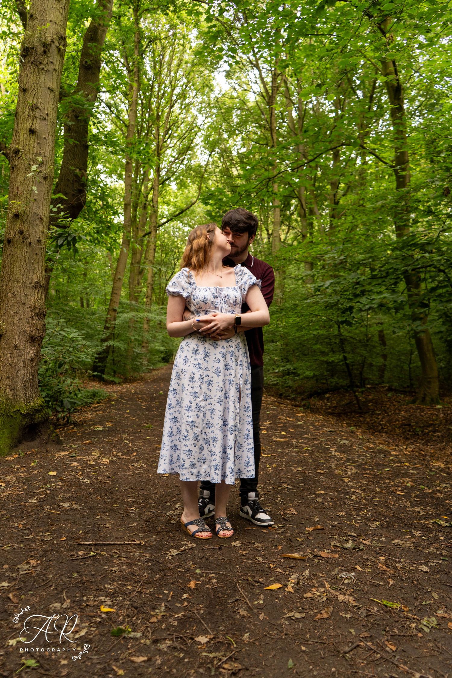 A couple embraces on a woodland path surrounded by tall trees and dappled sunlight.