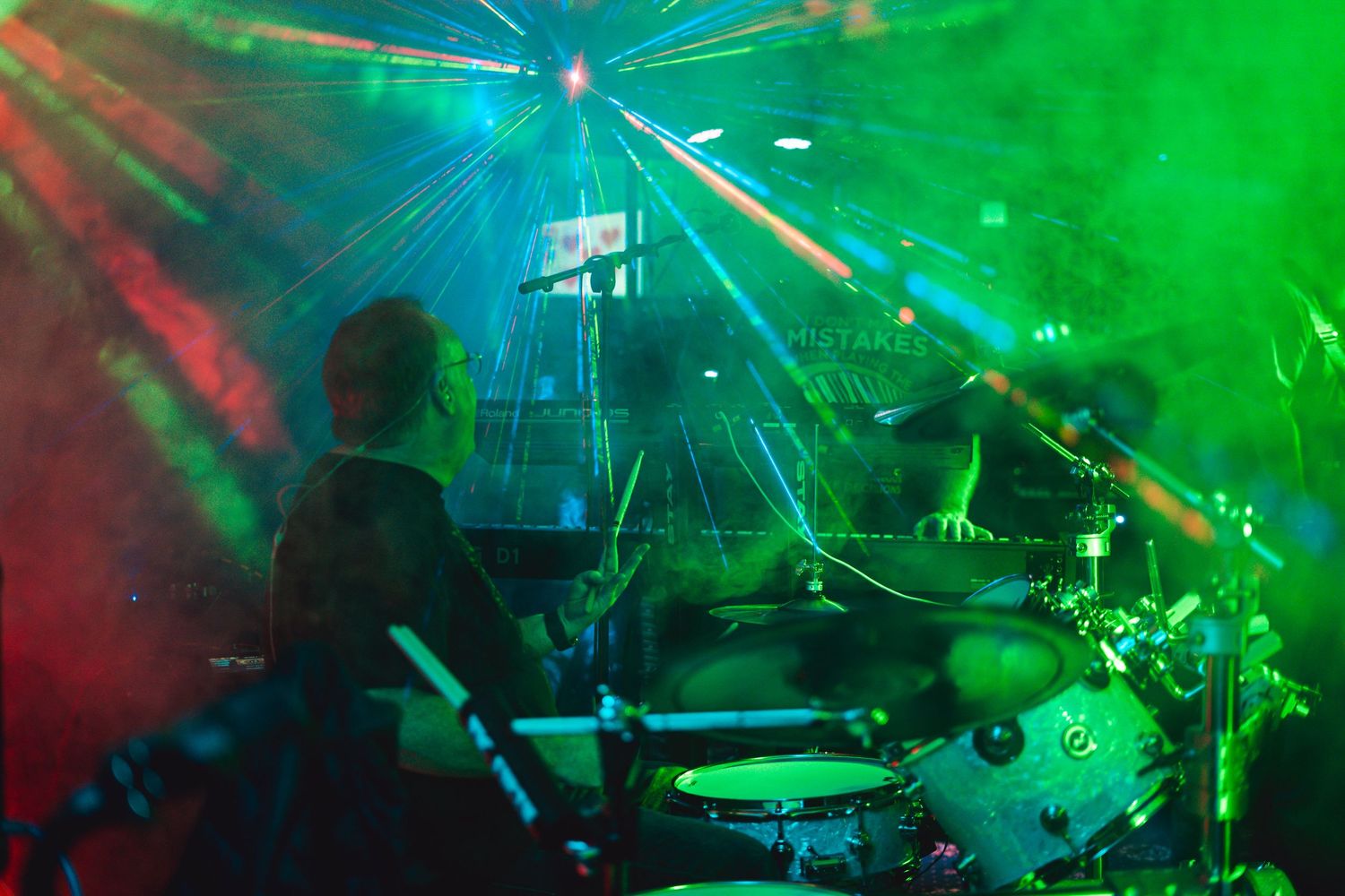 Drummer performing on stage with colorful green and red lighting effects.
