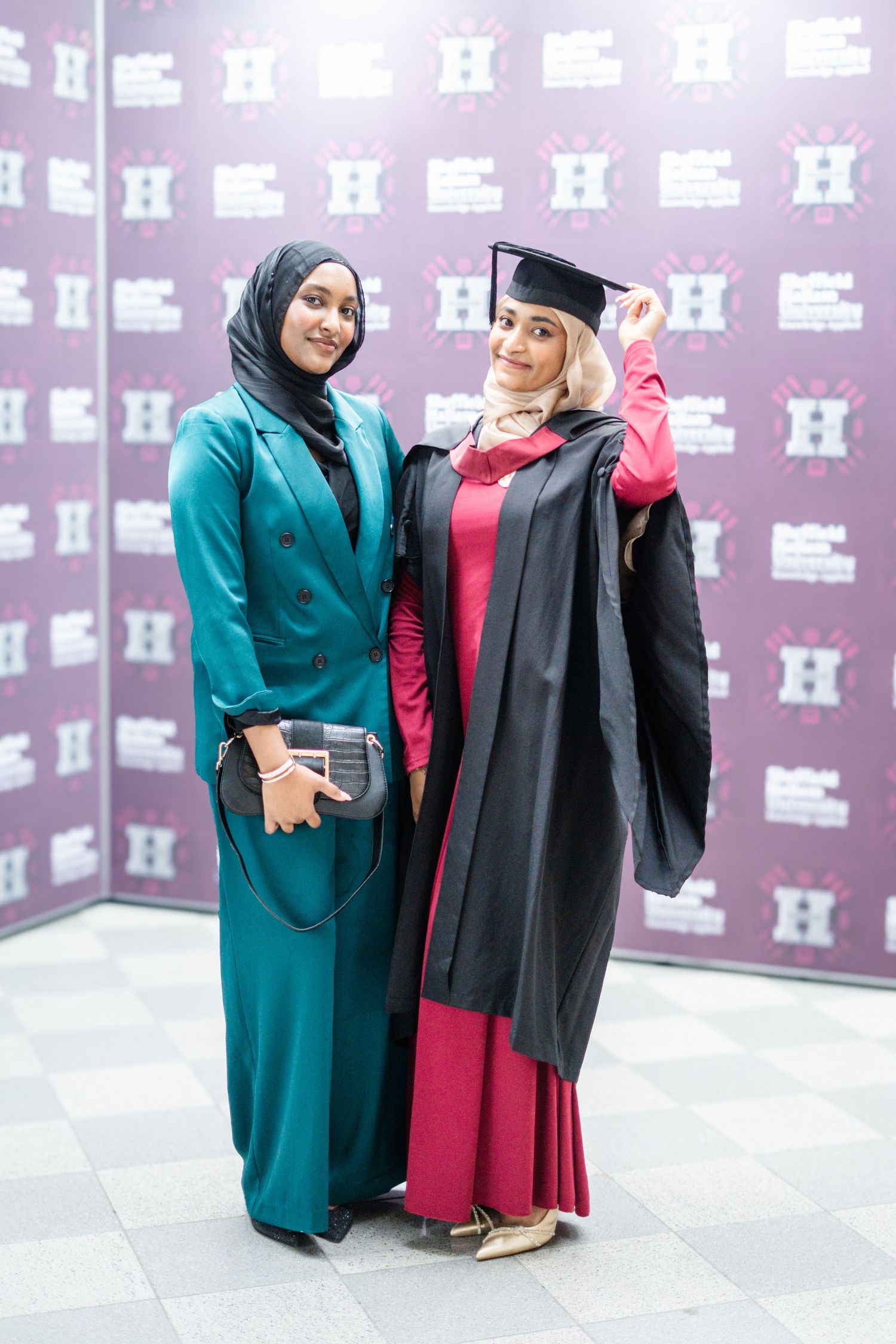 Two graduates in academic regalia celebrate together against purple patterned background.