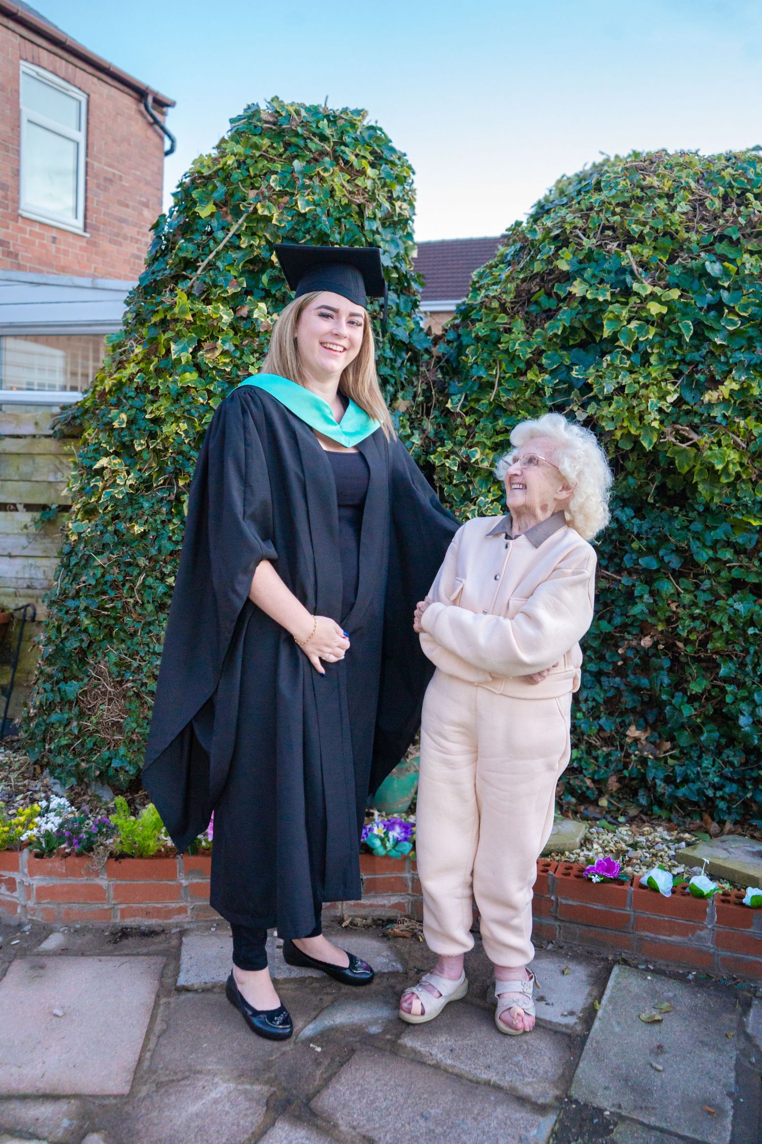 Graduate in academic regalia shares joyful moment with elderly loved one by brick wall and garden.