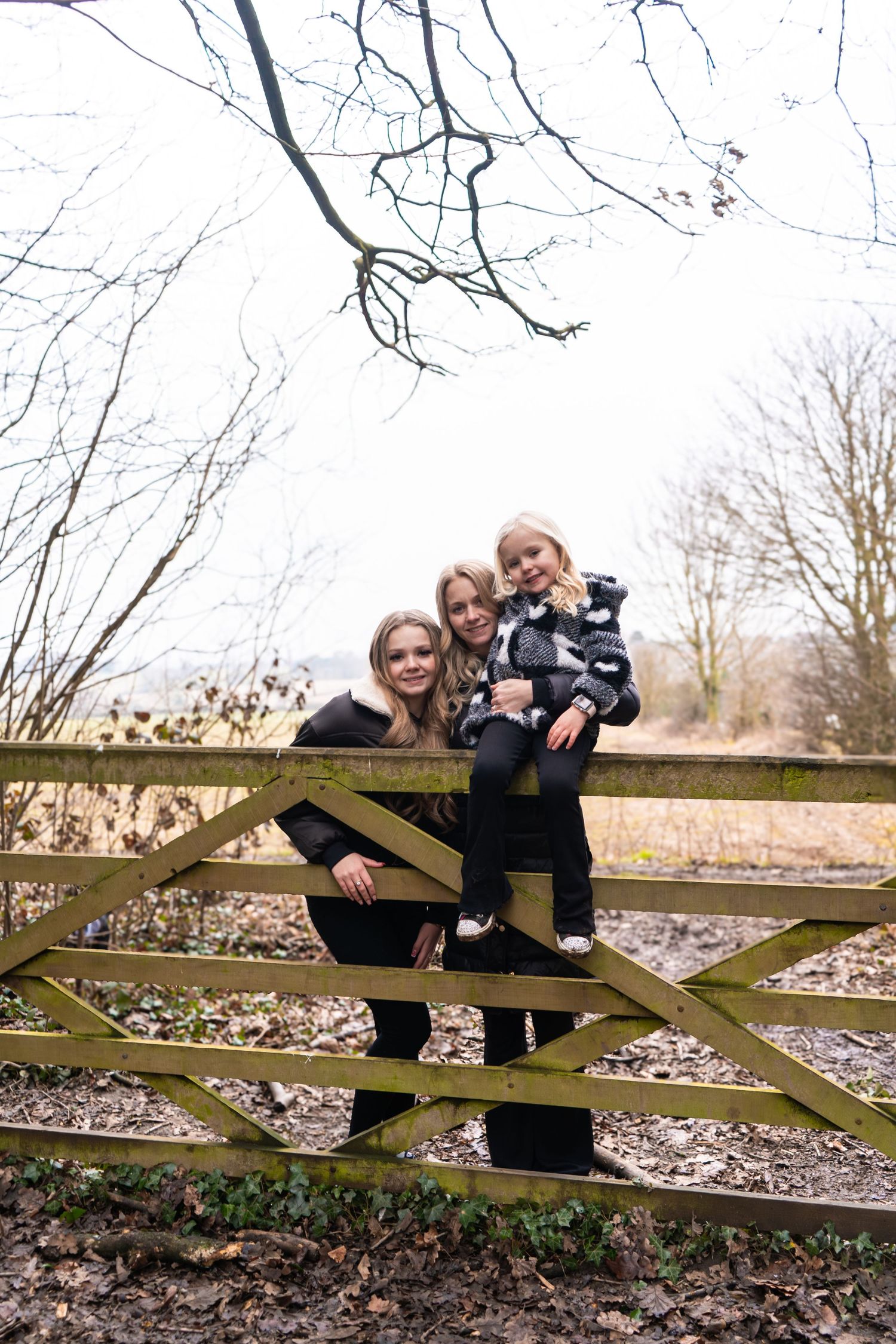 Three people pose on a wooden gate against a backdrop of bare winter trees.