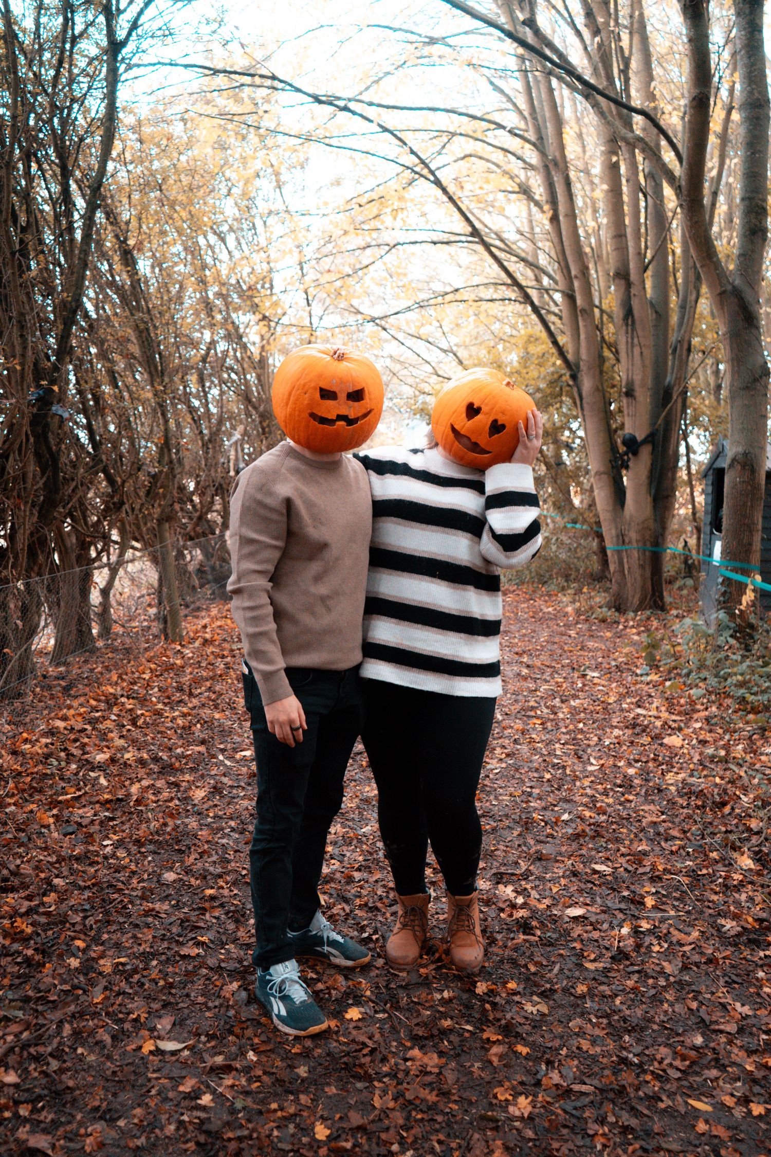 Two figures with jack-o-lantern heads stand together on an autumn forest trail.