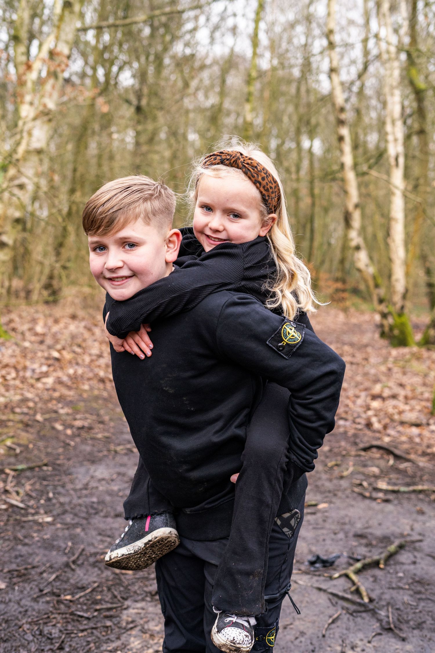 Playful siblings in matching black outfits hugging and laughing in a forest setting.