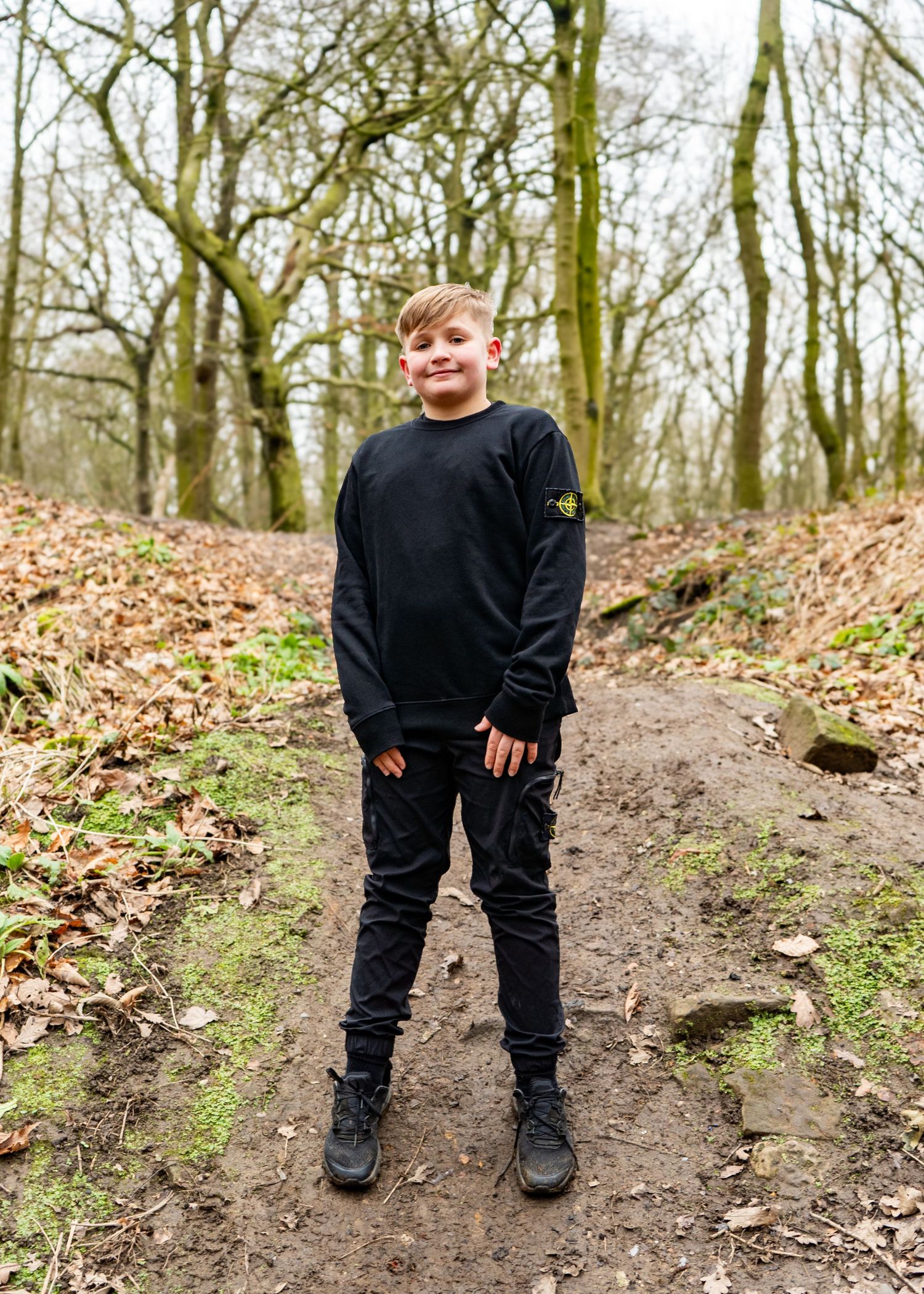 A child in all black clothing stands on a dirt path in a wooded area.