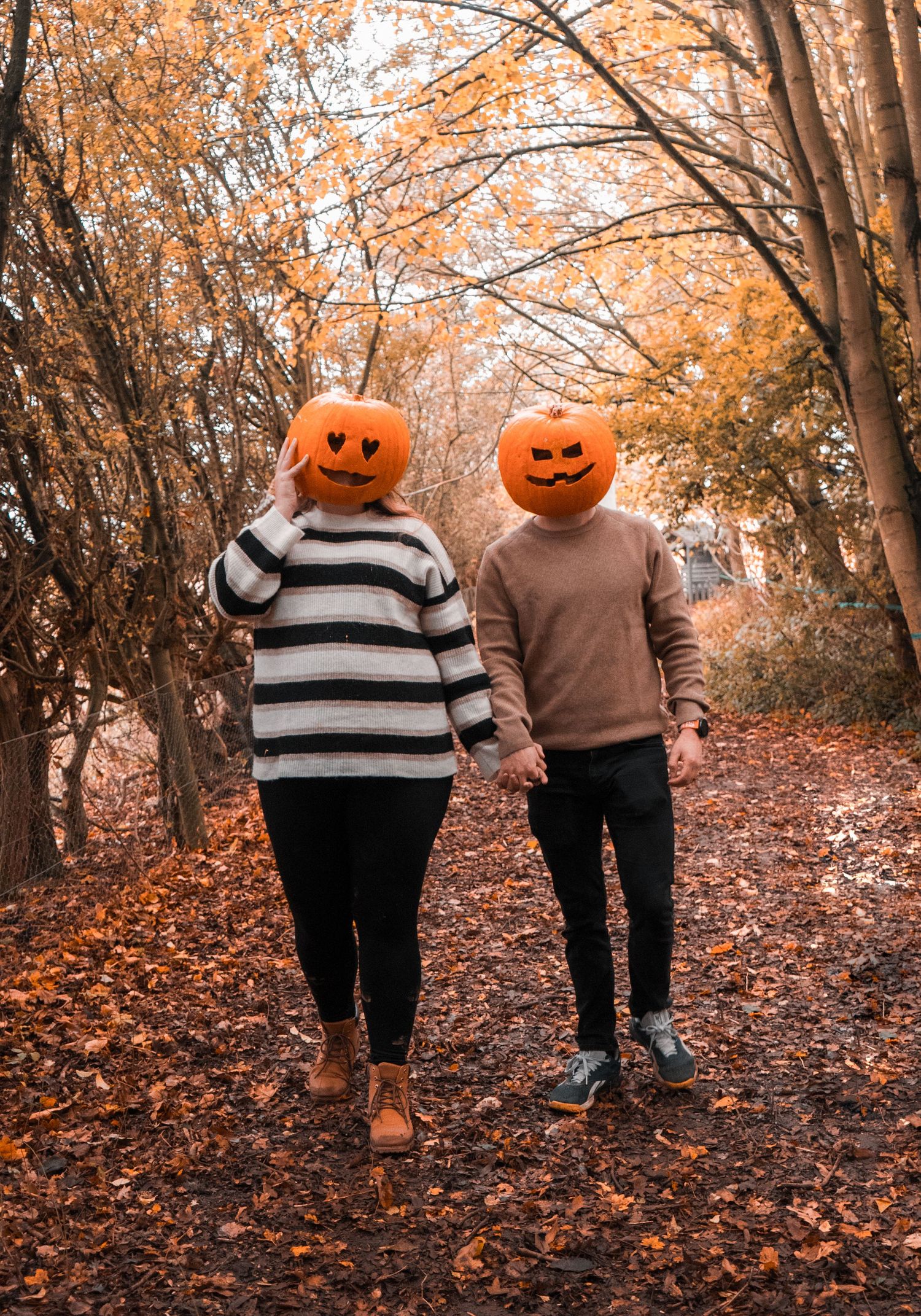 Two people wearing pumpkin heads walk together down a leaf-covered forest path in autumn.