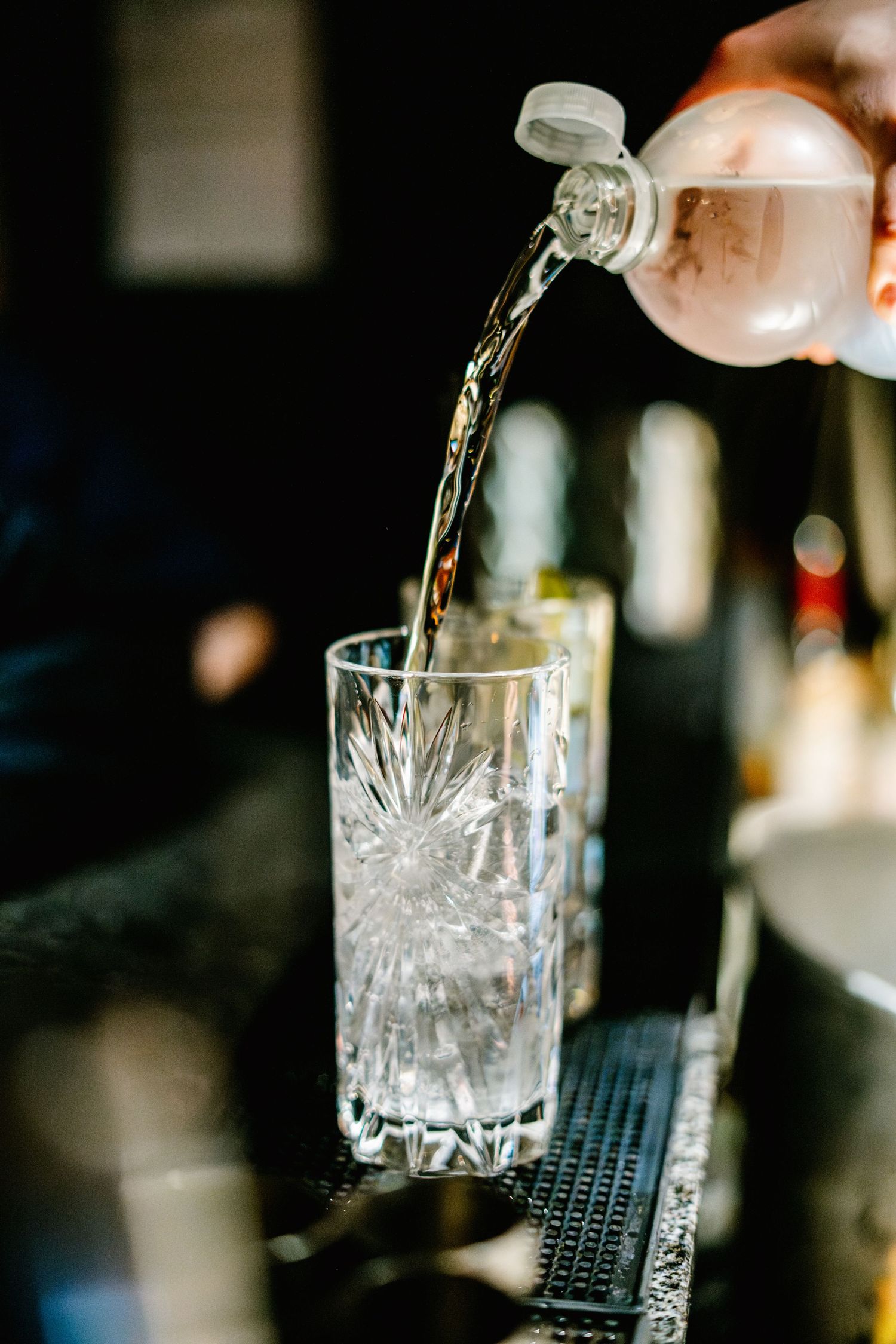 Crystal glass being filled with clear liquid from a bottle on a black bar mat.