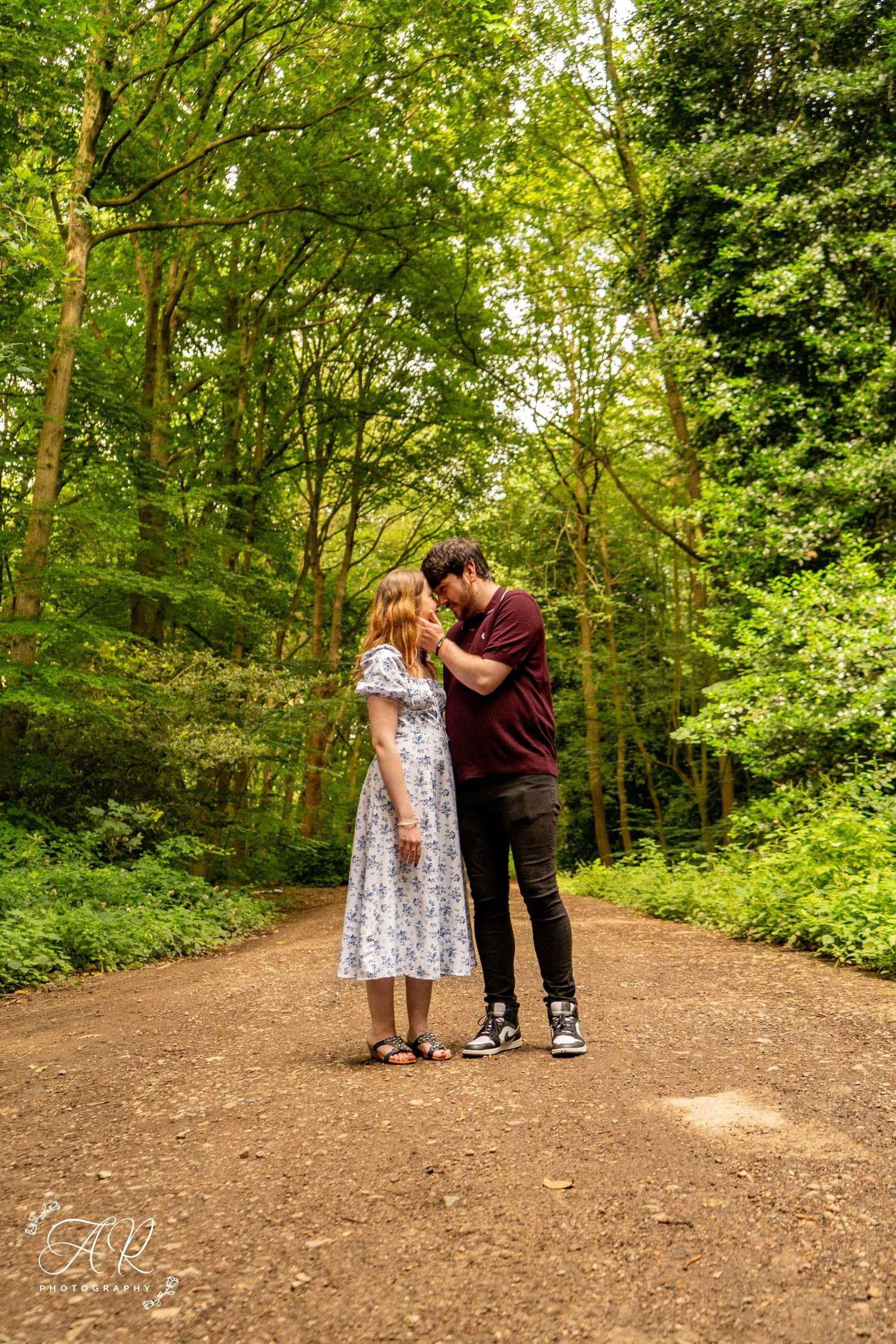 A couple shares an intimate moment on a forest trail surrounded by lush summer foliage.
