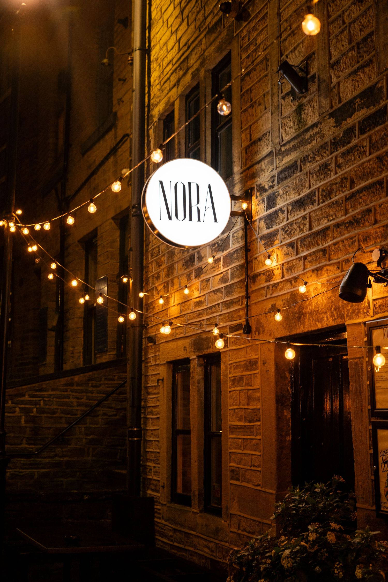 Brick building facade with illuminated NORA sign and string lights creating warm evening ambiance.