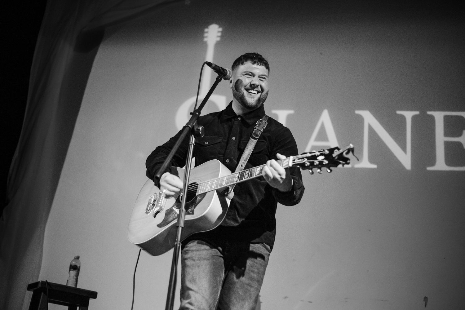 Black and white photo of performer playing acoustic guitar on stage with illuminated signage.
