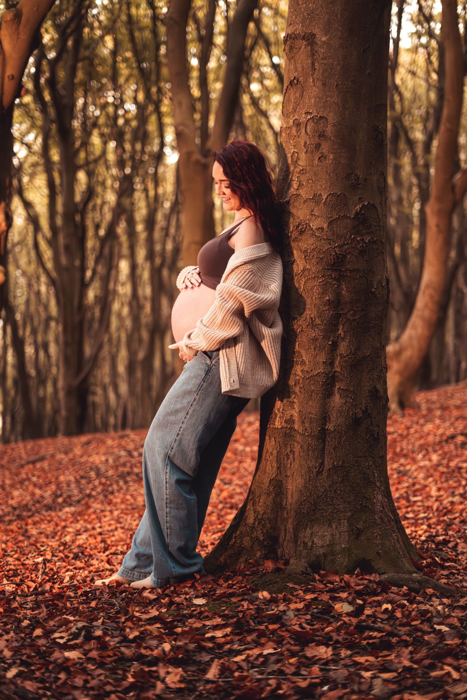 Person leaning against tree trunk in forest covered with orange autumn leaves.