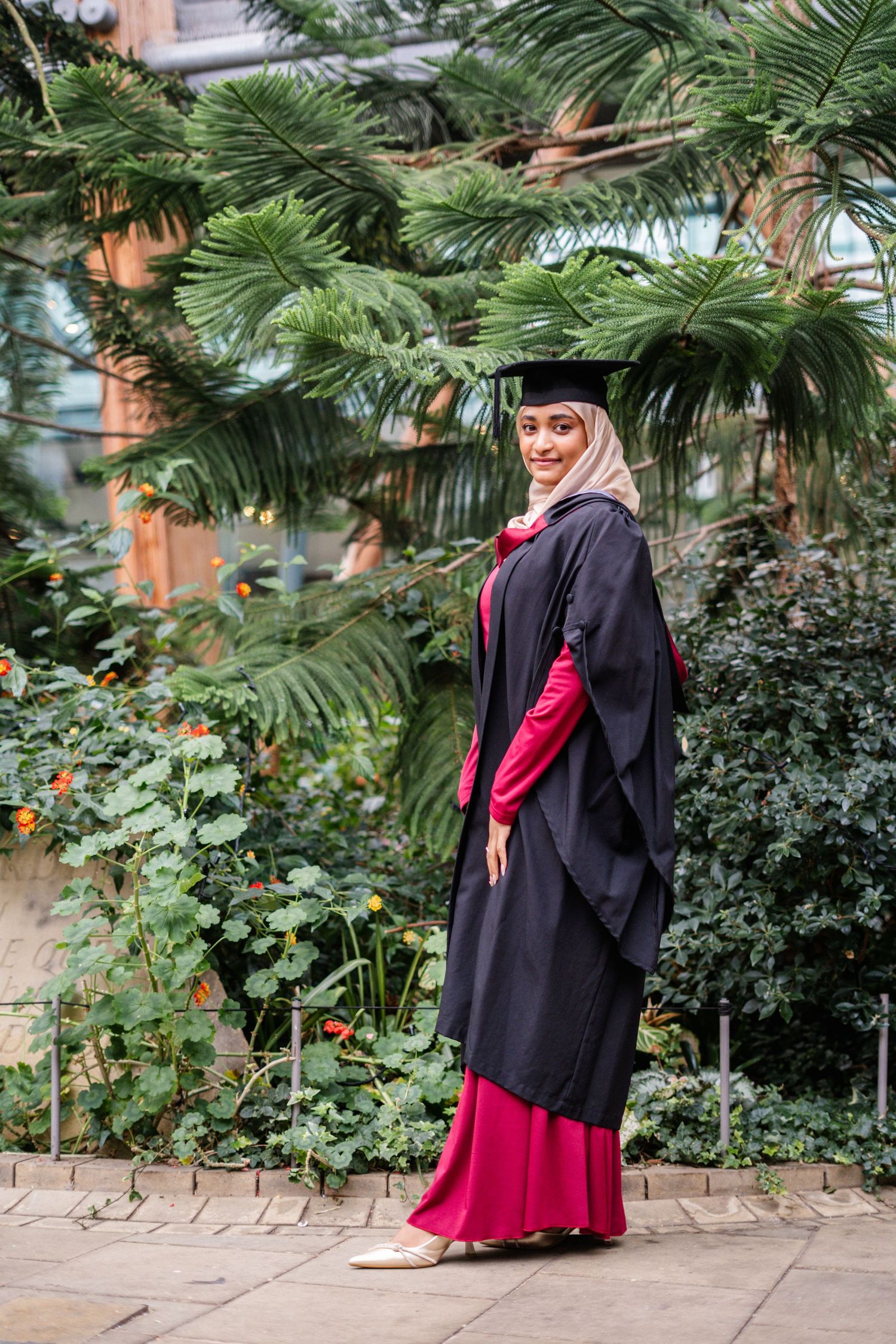 Graduate wearing black academic regalia and red dress poses near palm trees on campus.
