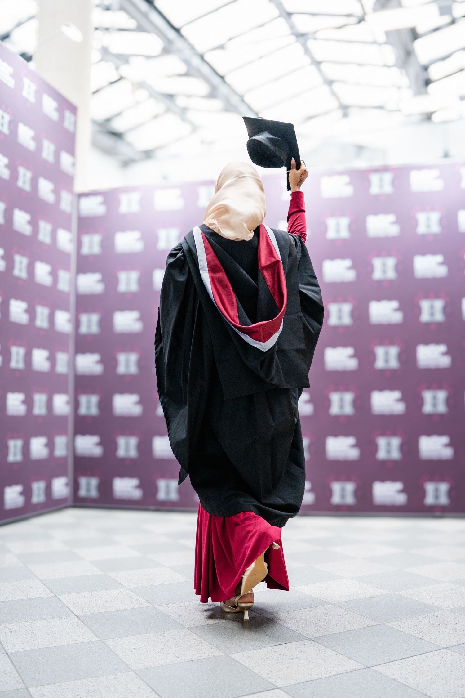 Graduate in black and red academic regalia poses gracefully on tiled floor with geometric backdrop.