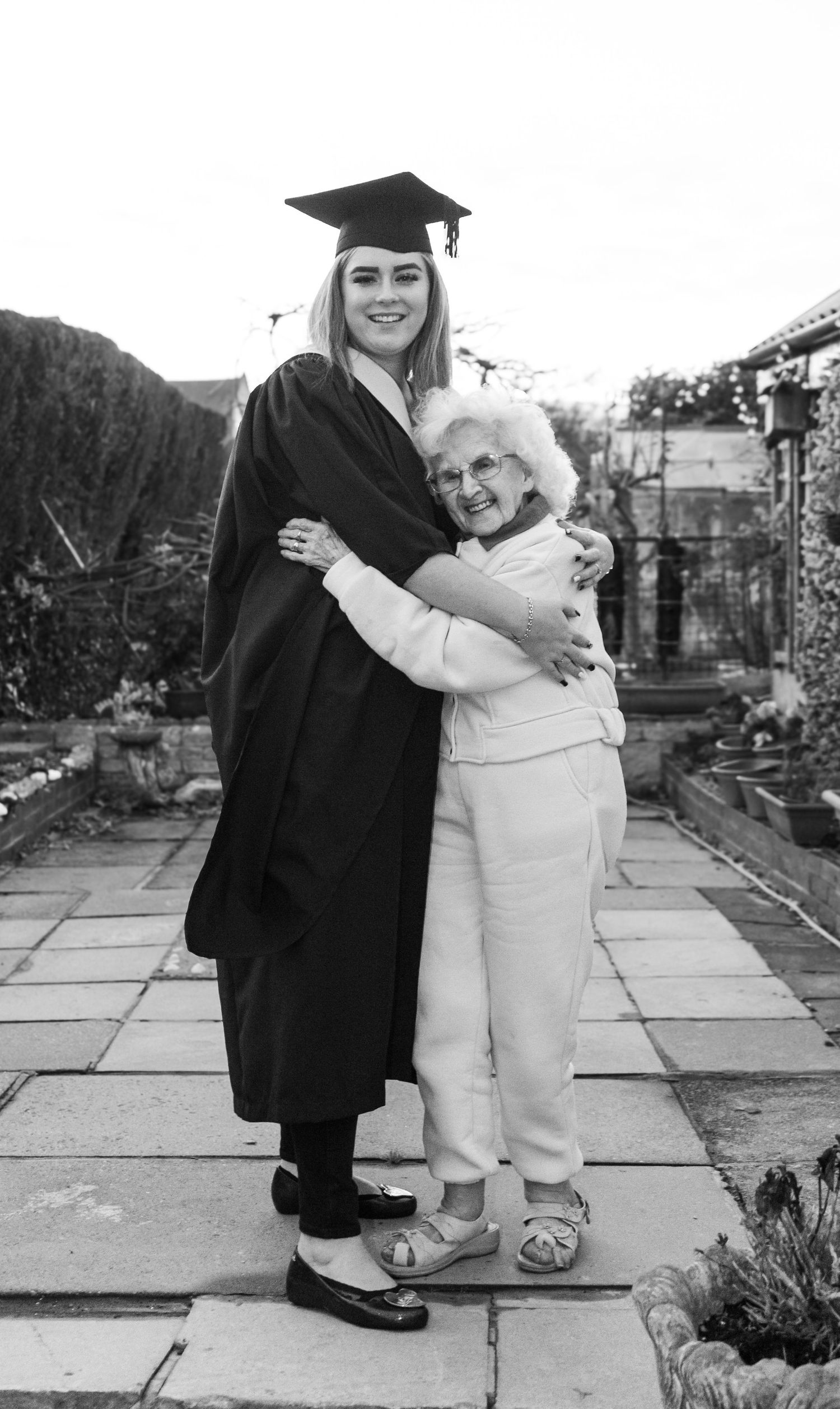 Black and white graduation photos showing graduate and elderly relative embracing on stone patio.