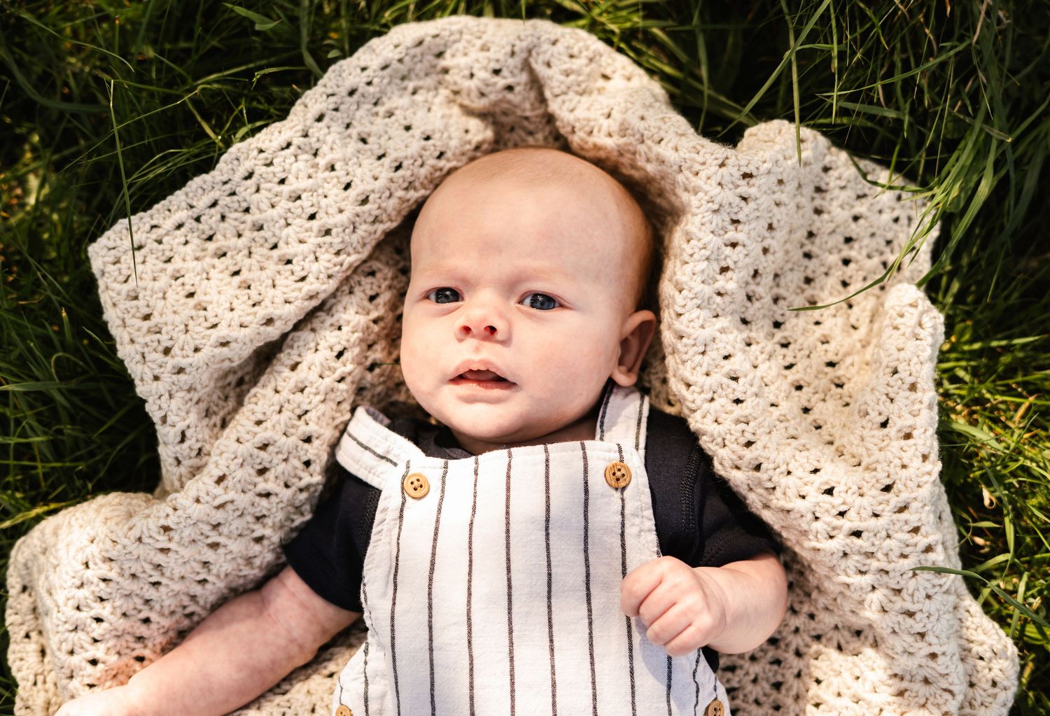 Baby resting on cream colored knit blanket in grass during outdoor photo session.