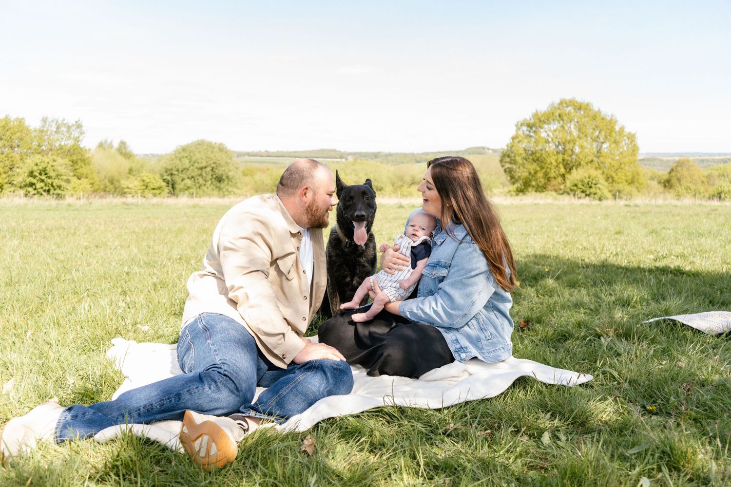 Family relaxing on blanket in sunlit meadow with rolling hills in background.