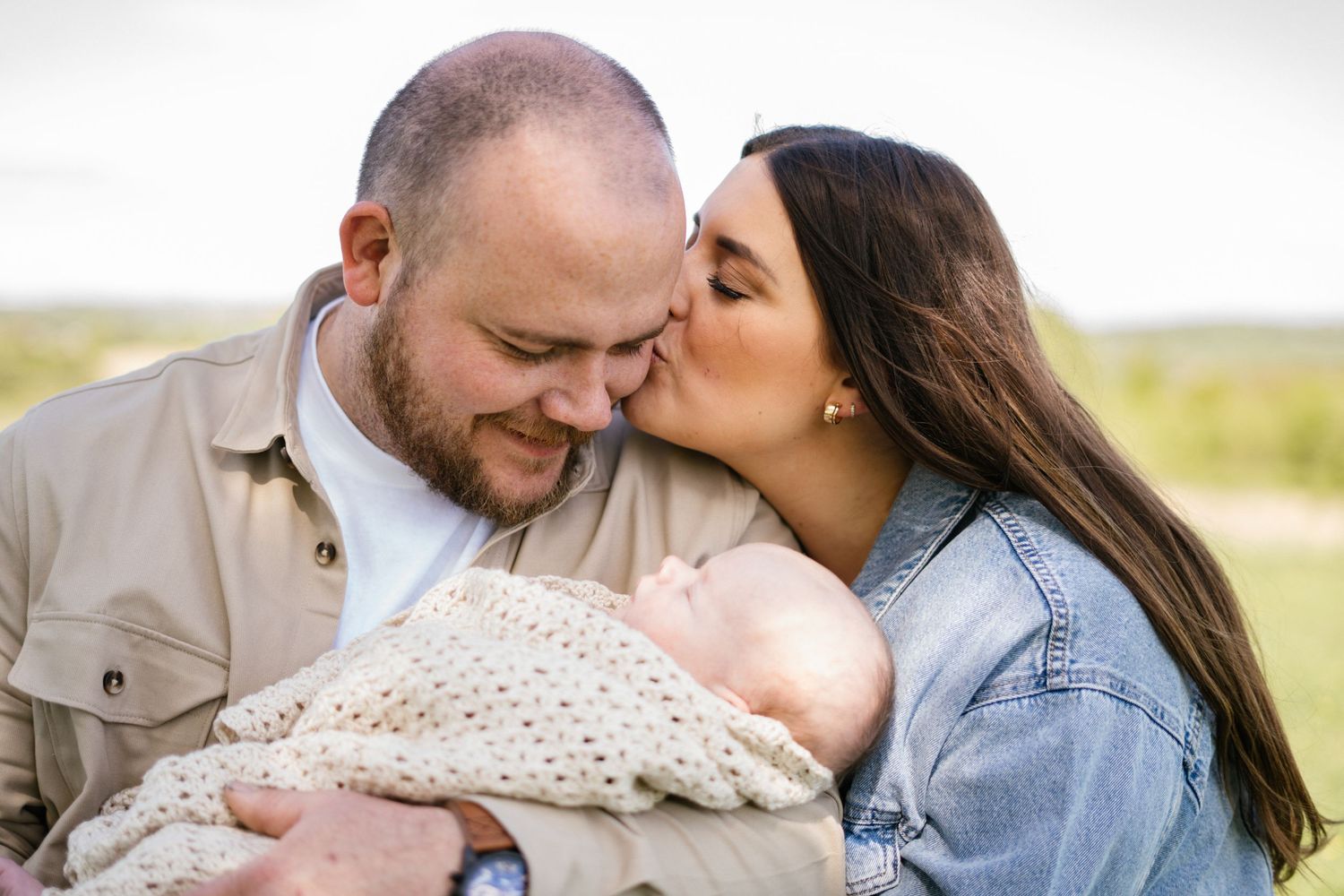 Couple sharing intimate moment on grassy field while holding newborn in cream blanket.