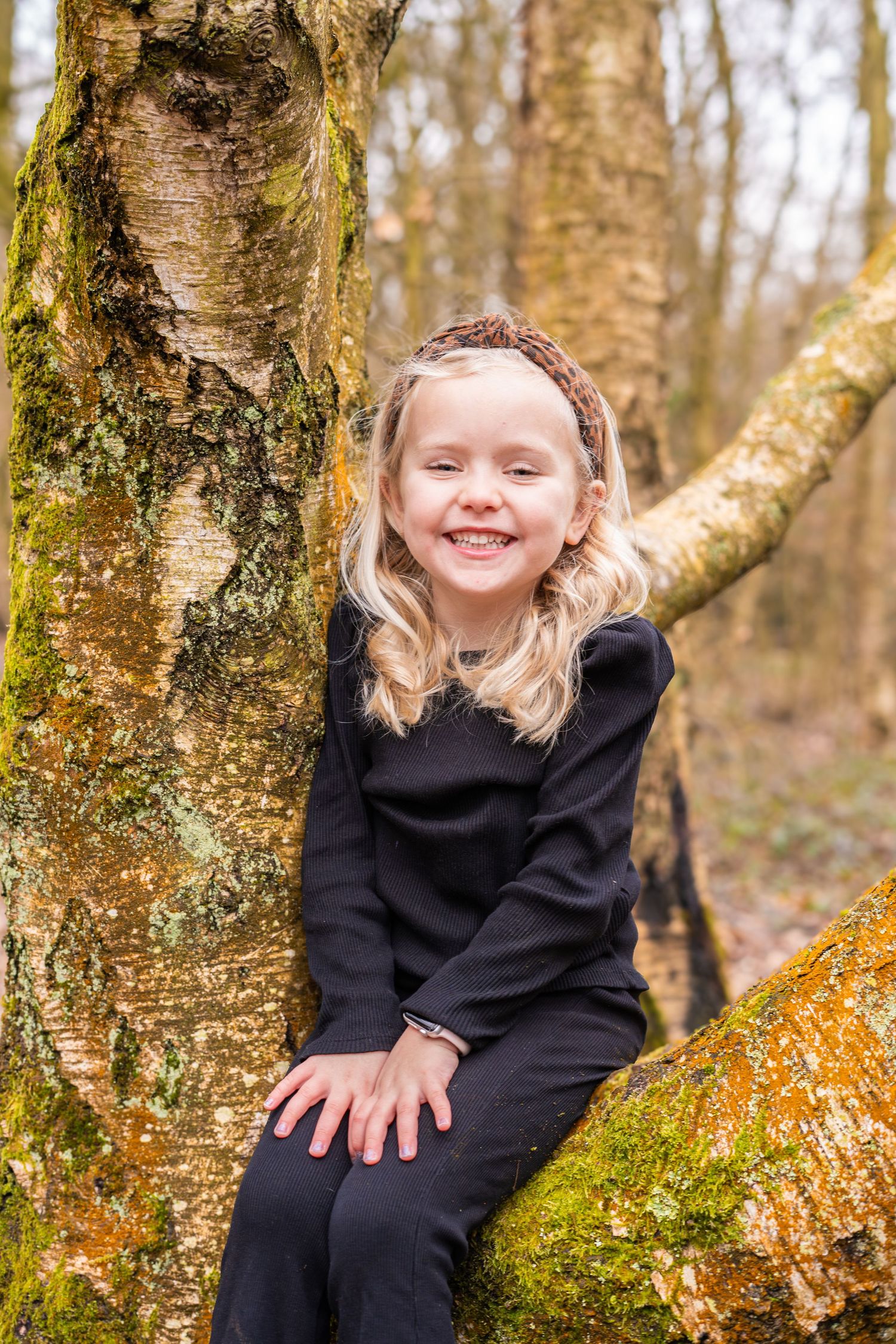A child in a black sweater sits on a mossy tree branch in an autumn forest setting.