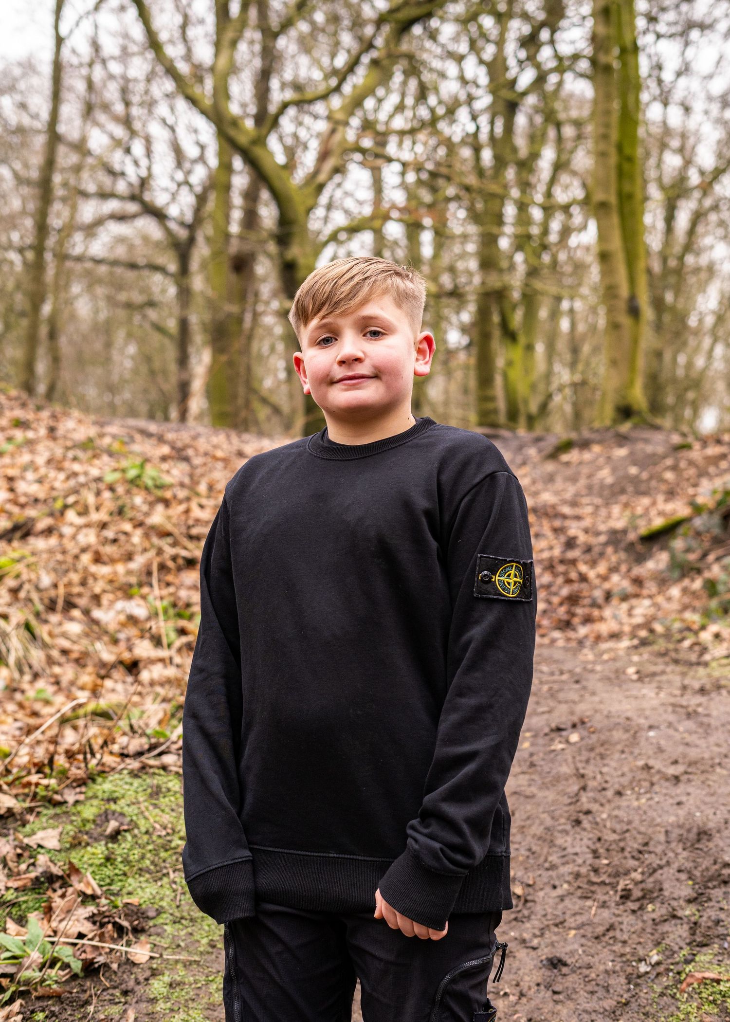 A child wearing a black sweater stands on a forest trail surrounded by trees.