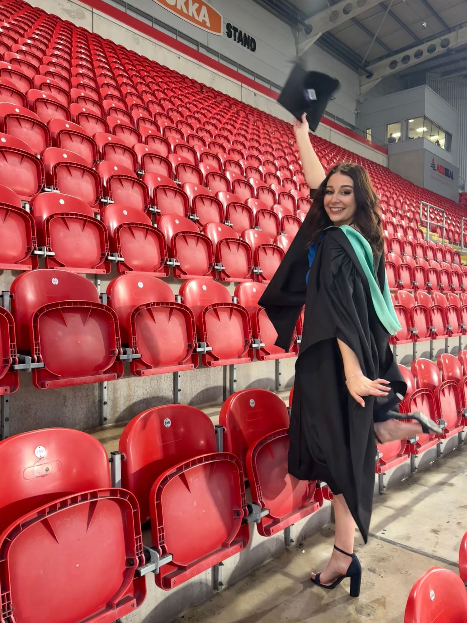 Graduate in black cap and gown poses in front of red stadium seats during graduation celebration.