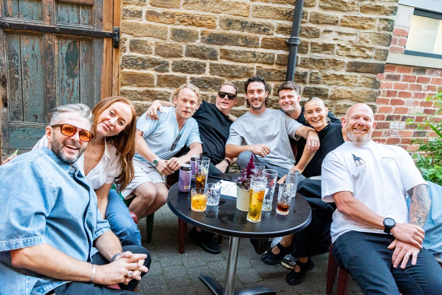 Group enjoying drinks at outdoor patio table with stone wall backdrop.