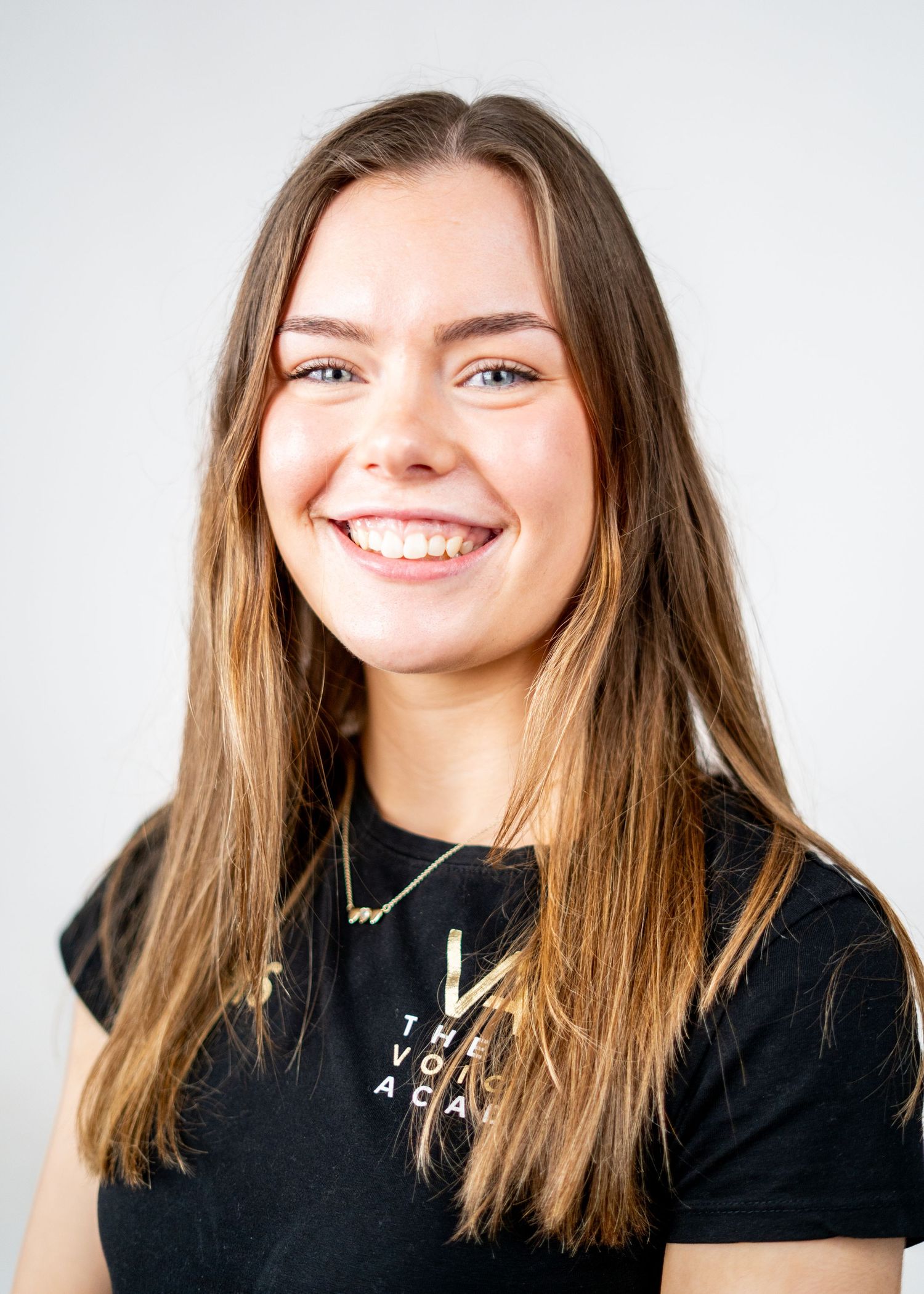 Professional headshot of a person in a black t-shirt with long brown hair against a white background.