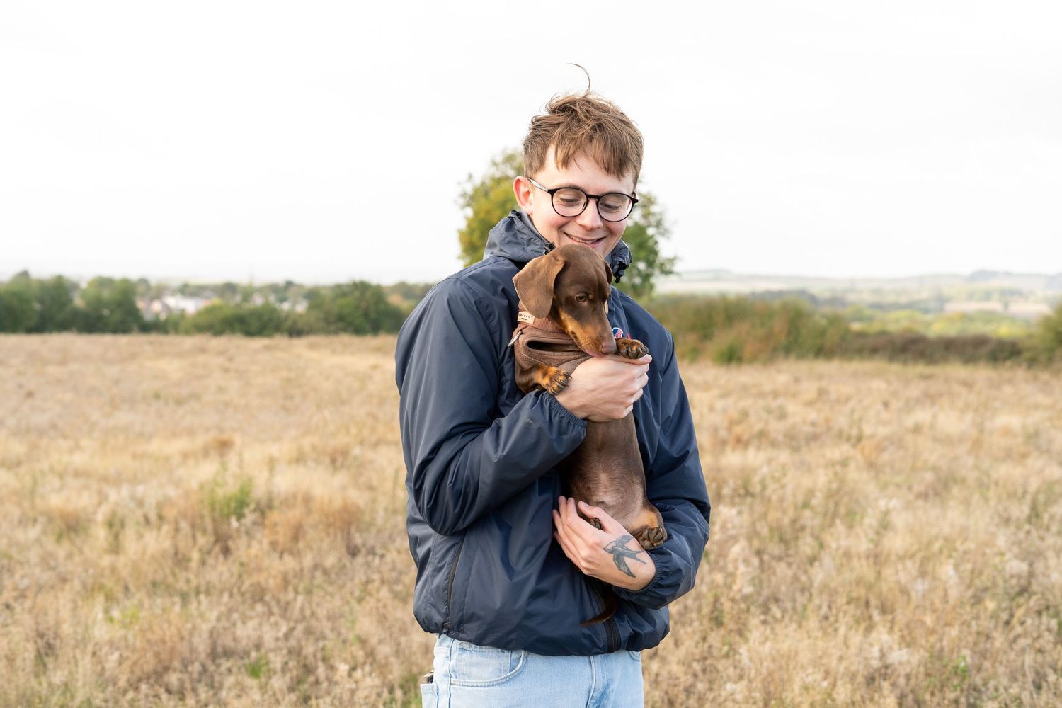 A sweet dachshund puppy being embraced in a navy jacket against an autumn field backdrop.