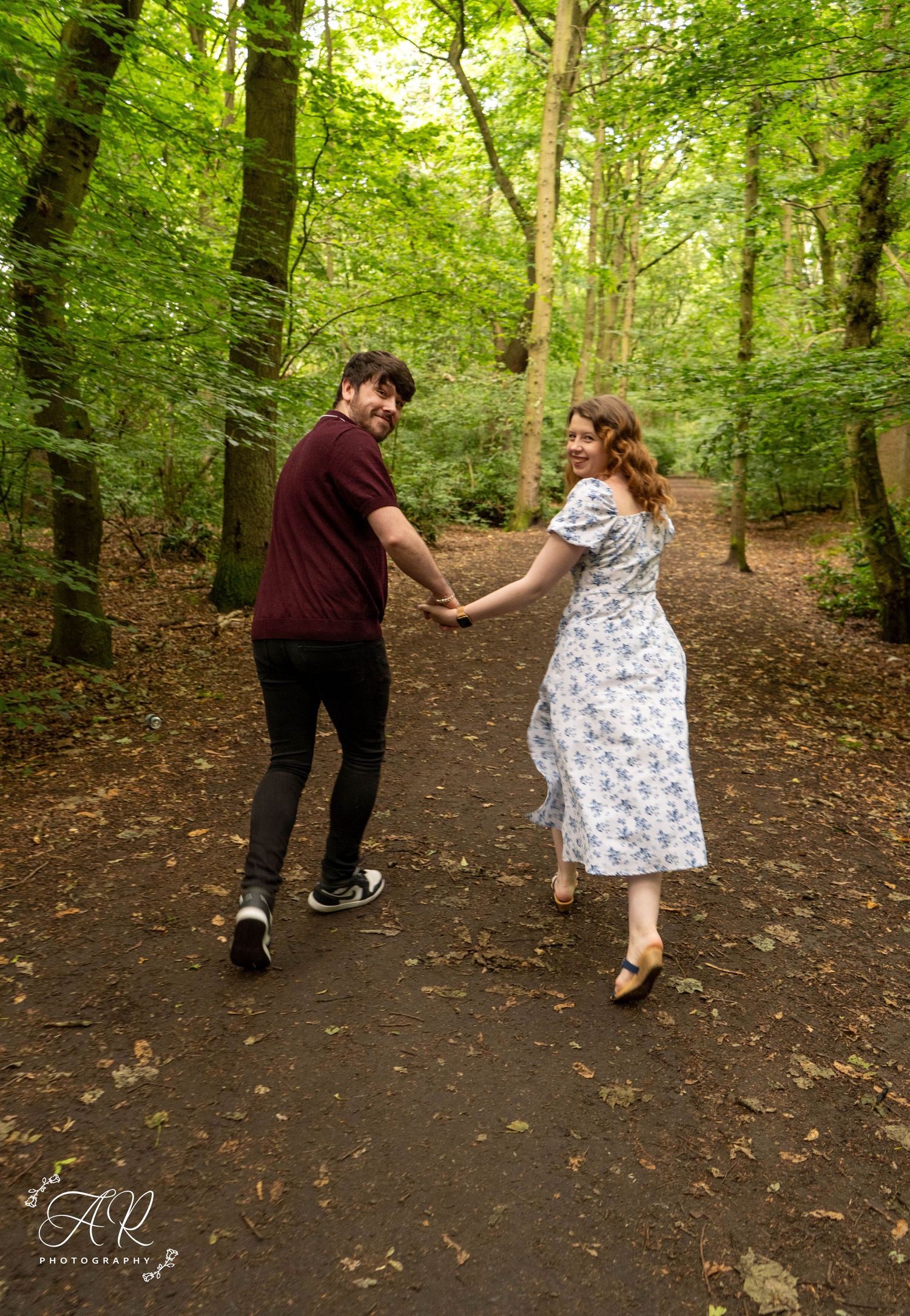 A couple dances playfully along a forest trail surrounded by green trees.