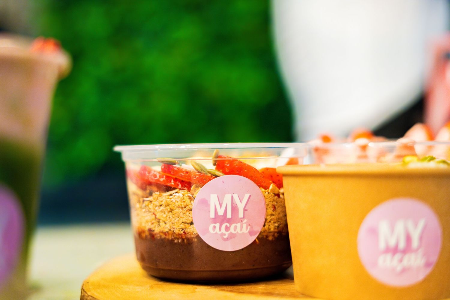 Colorful food bowls with pink branding displayed on a wooden surface with green foliage background.