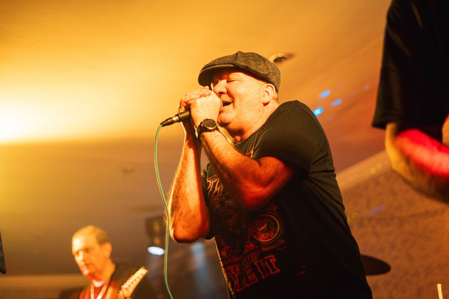 Performer in black t-shirt and cap sings passionately into microphone against golden background lighting.