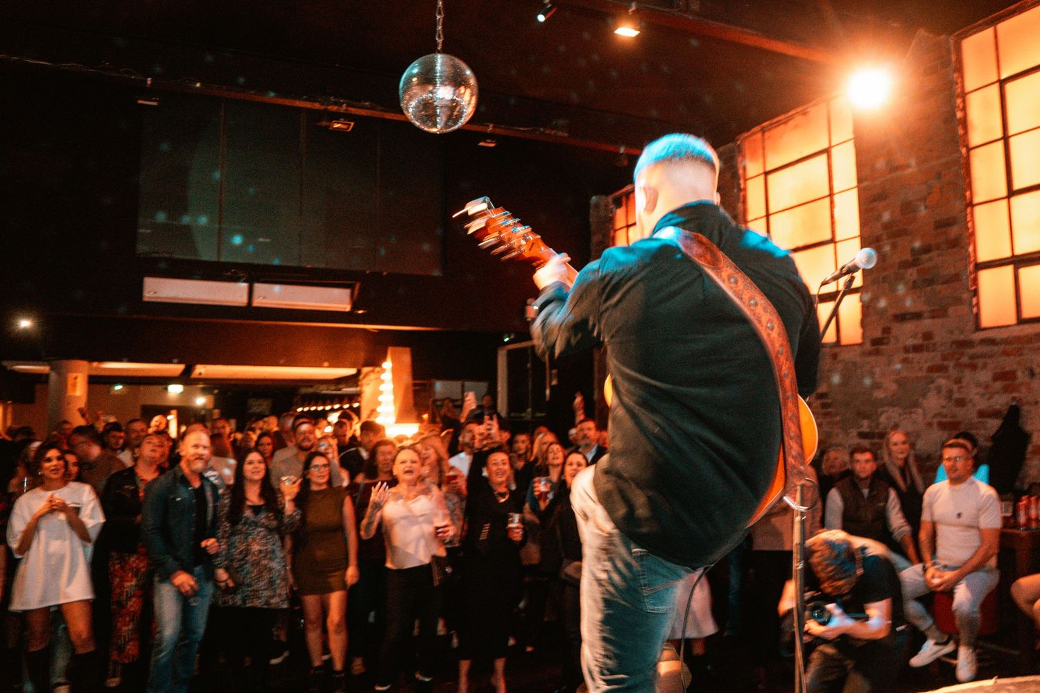 Musician performs on stage with disco ball and crowd at energetic concert venue.