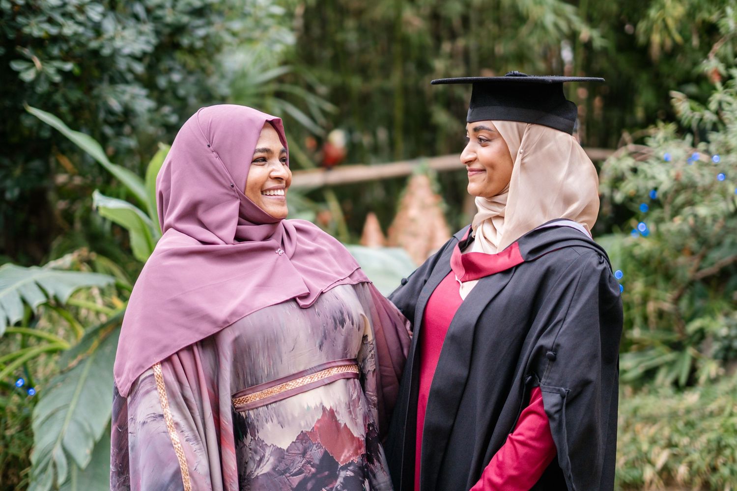 Graduates wearing black gowns and hijabs celebrate together in an outdoor garden setting.