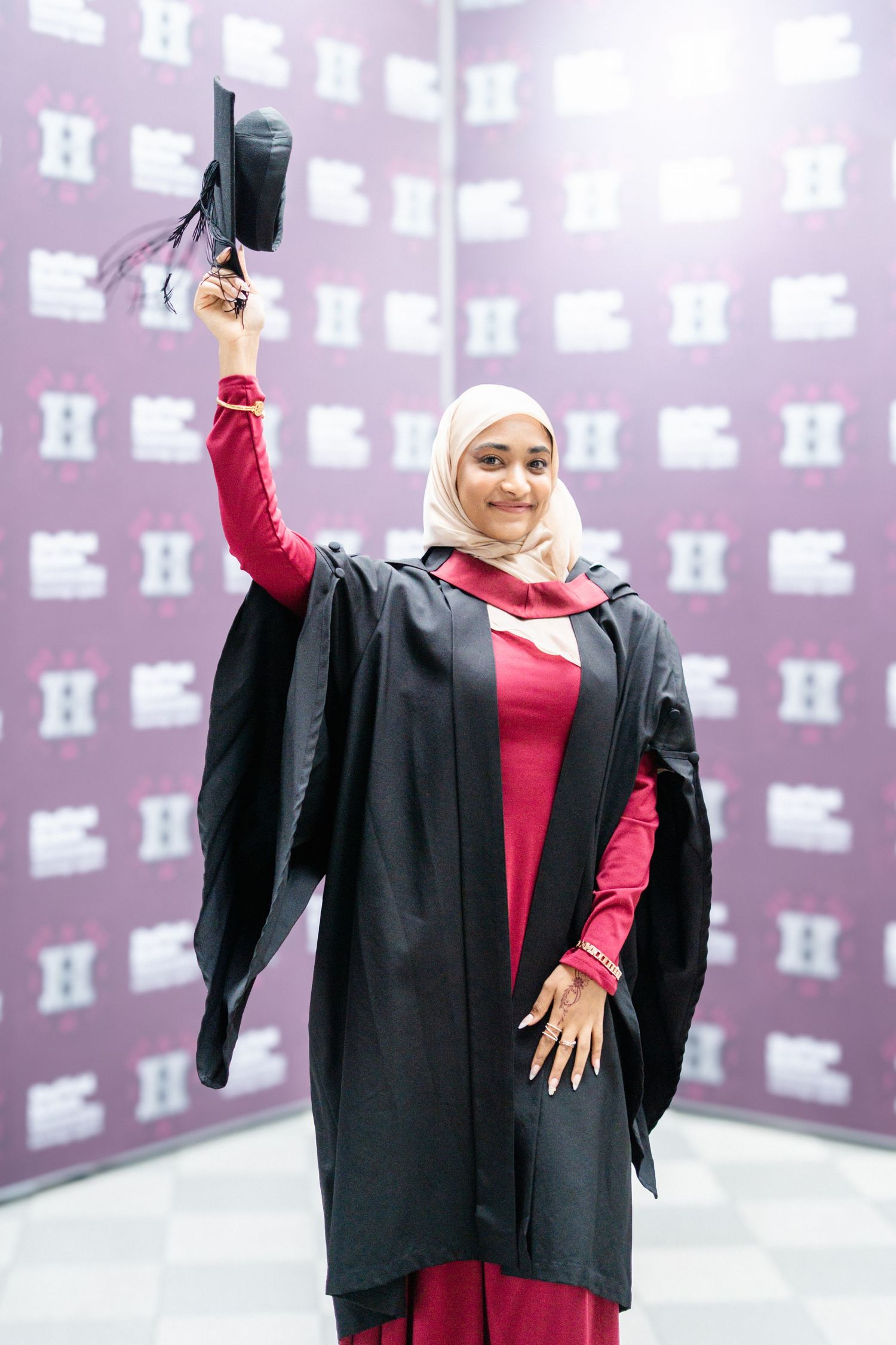 Graduate celebrating with diploma in traditional black gown and red dress against patterned wall.