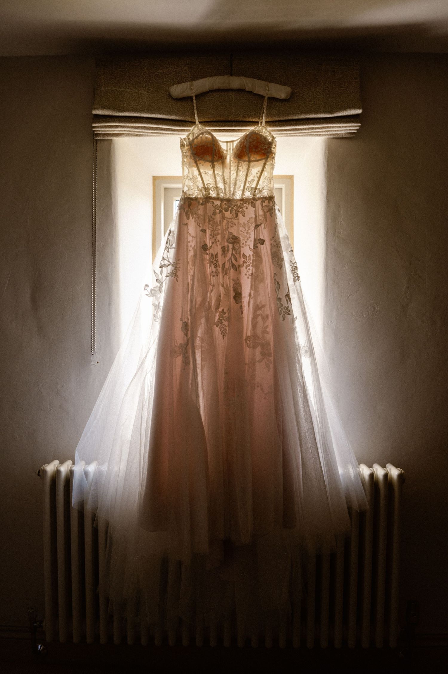 Ethereal wedding dress backlit by window light in a dark room.