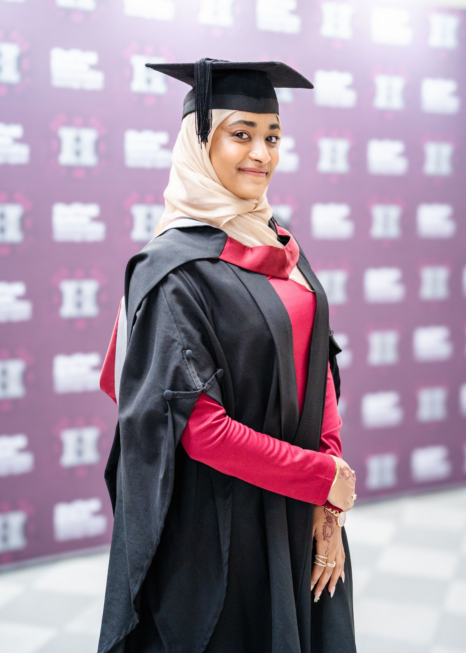 Graduate in traditional academic dress and hijab poses against decorative purple wall.